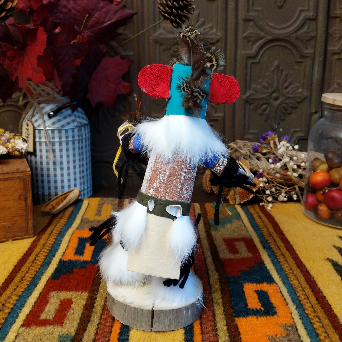 This colorful Navajo Quail Kachina by Debbie James features a turquoise hand-painted bird mask with red accents, a yellow beak, natural feather crest, white fur, leather ties, and a wooden display base; back view.