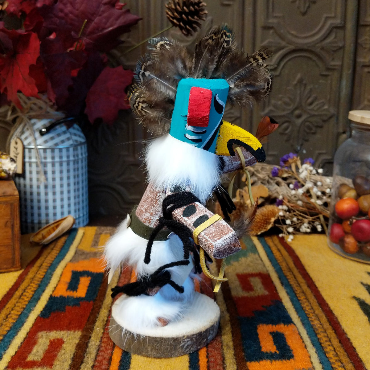 This colorful Navajo Quail Kachina by Debbie James features a turquoise hand-painted bird mask with red accents, a yellow beak, natural feather crest, white fur, leather ties, and a wooden display base; side view.