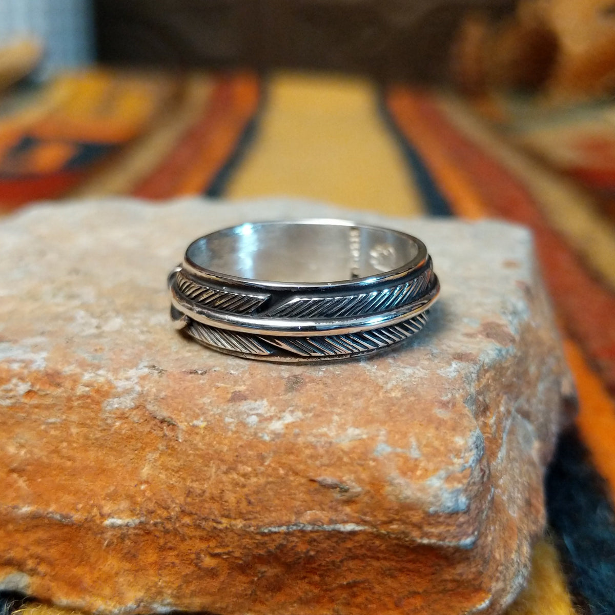 Silver feather-patterned band ring featuring diagonal engraved lines, darkened recesses, and a raised polished silver ridge around the center.