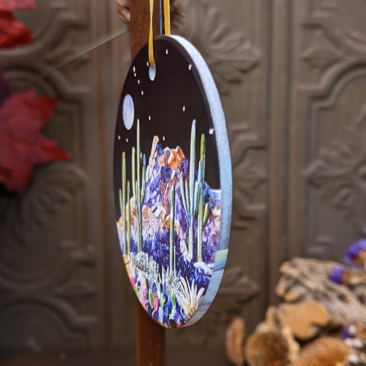An oval hanging ornament with a hand-painted desert night scene, featuring purple and green cacti, a mountain, and a full moon against a dark sky, suspended by a yellow string.