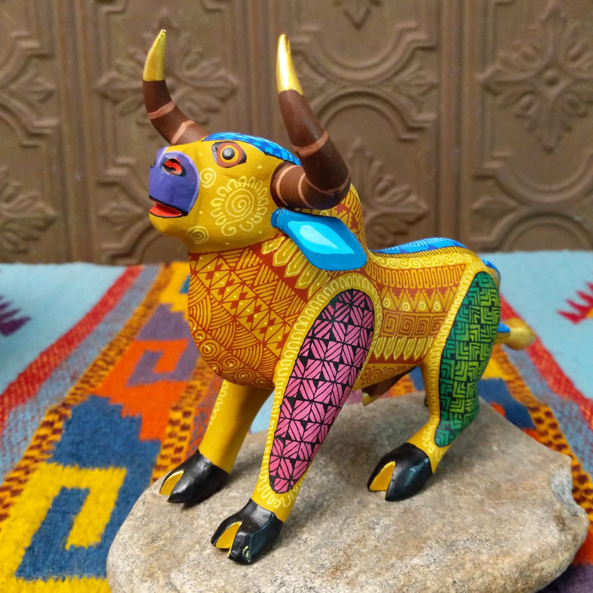 Oaxacan bull wood carving by Abundio Muñoz with curved horns, a golden yellow body, and colorful geometric patterns across the legs and torso.