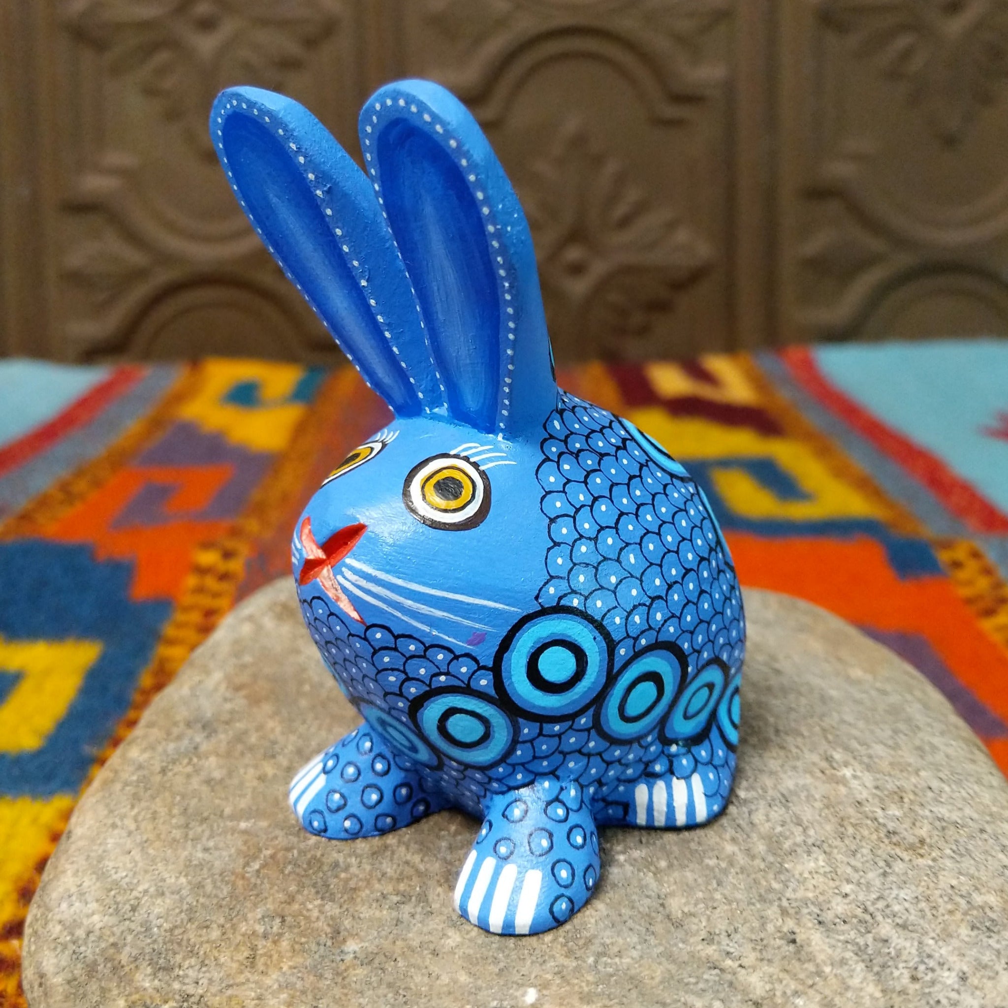 Blue Oaxacan bunny wood carving by Alberto Hernandez with tall ears, ringed eyes, and layered circular and scale-like painted patterns.