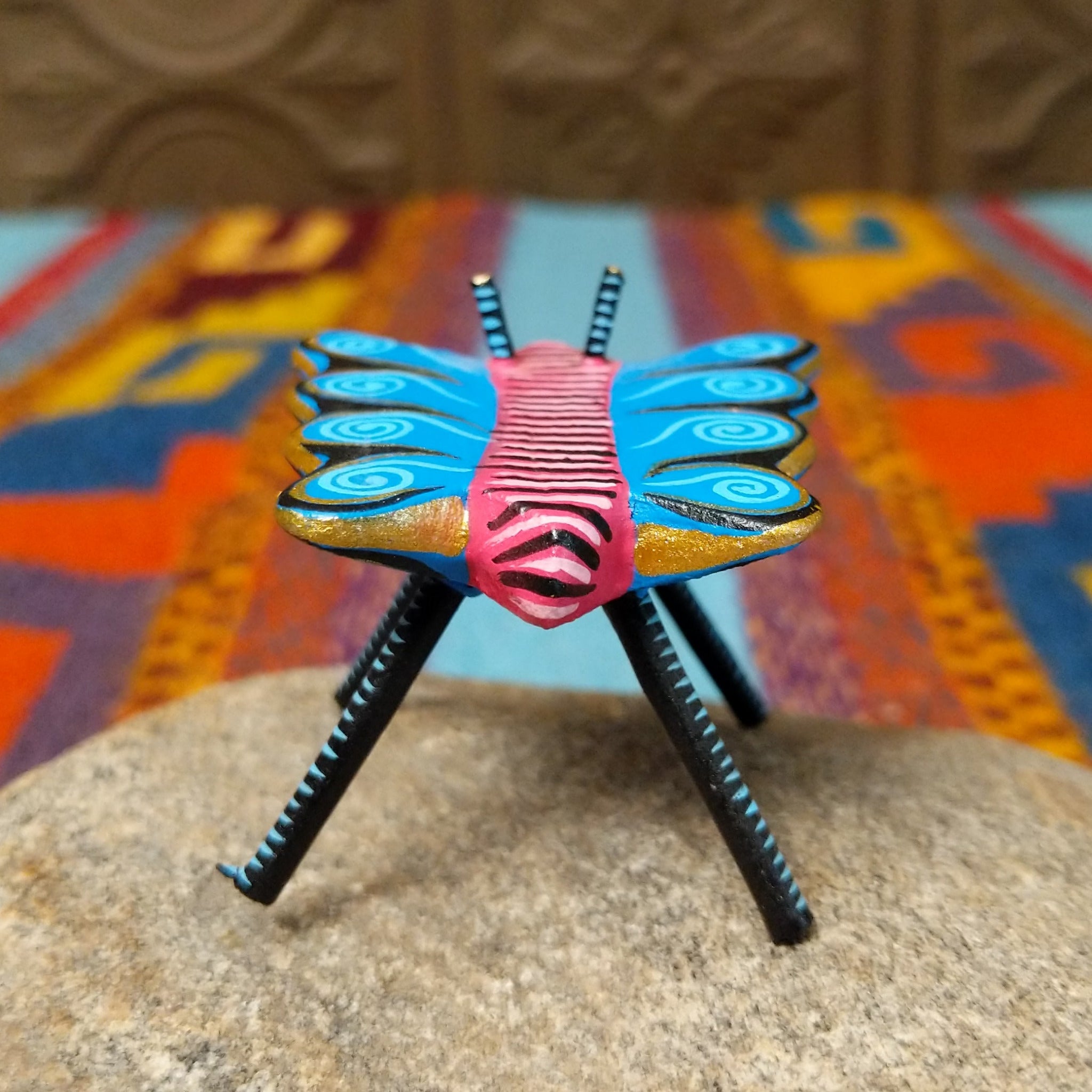 This colorful Oaxacan butterfly wood carving features a pink body, turquoise wings with spiral patterns, gold accents, striped antennae, and long black-and-turquoise legs; rear view.
