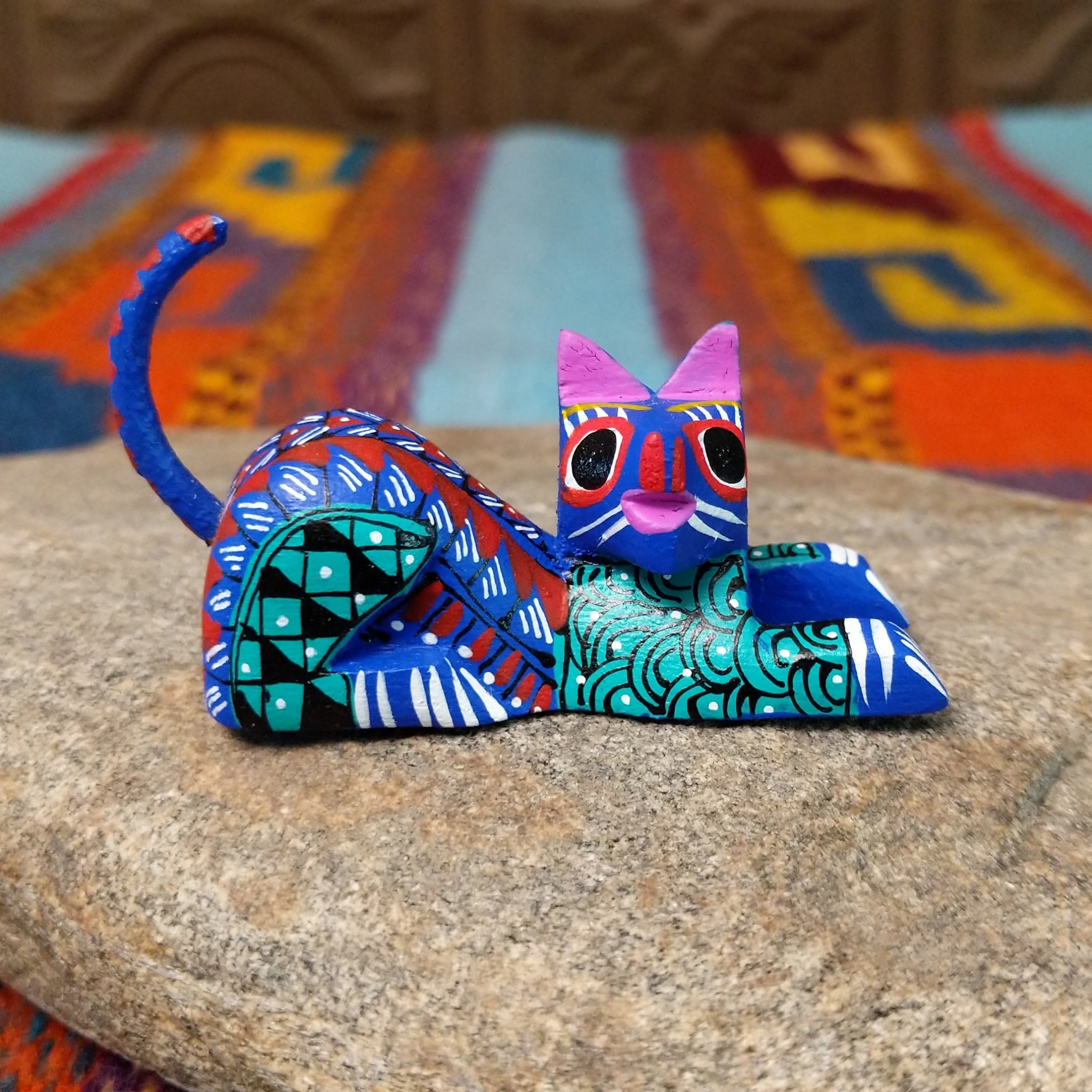Reclining Oaxacan cat wood carving with a curled tail, oversized eyes, and colorful hand-painted geometric patterns.
