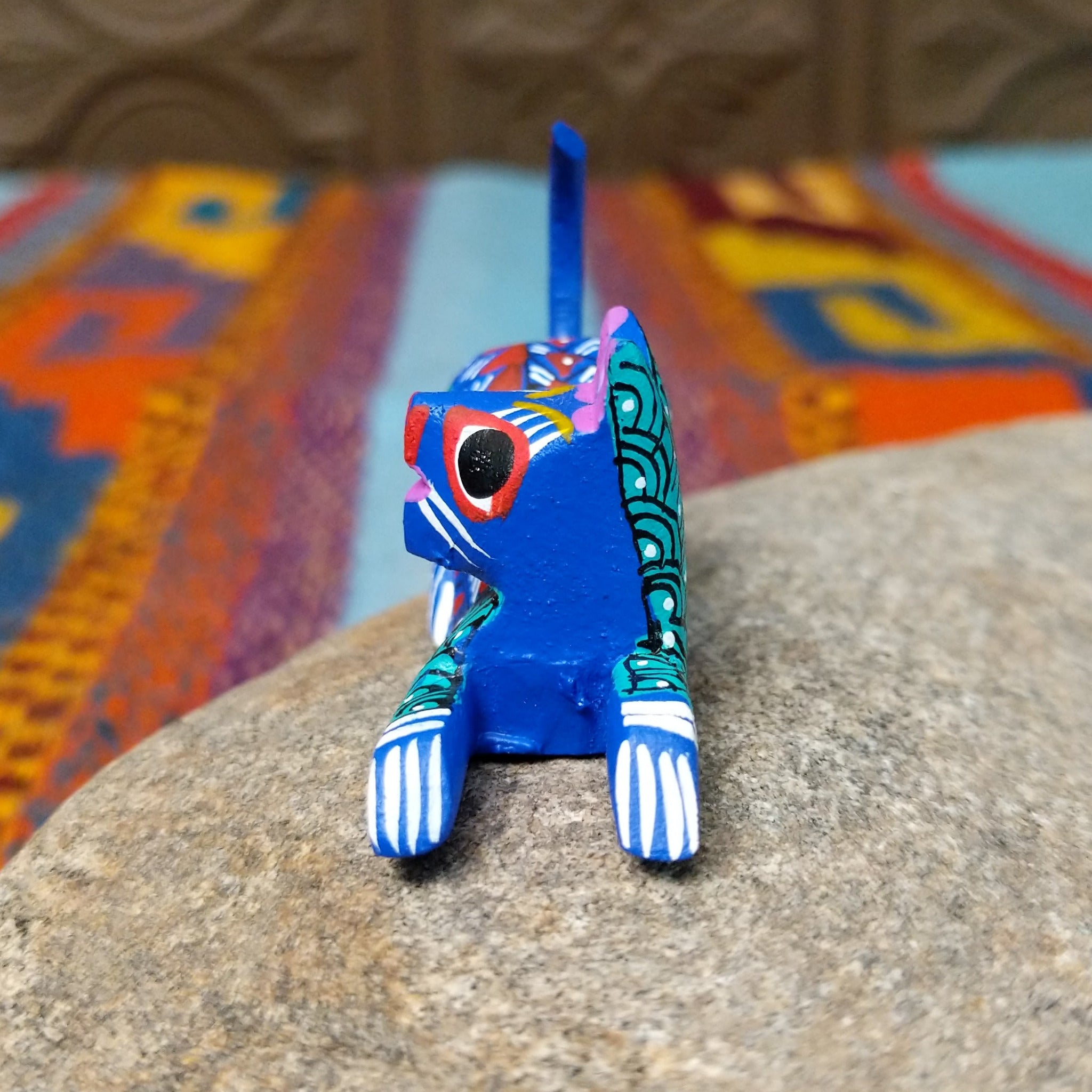 Reclining Oaxacan cat wood carving with a curled tail, oversized eyes, and colorful hand-painted geometric patterns.
