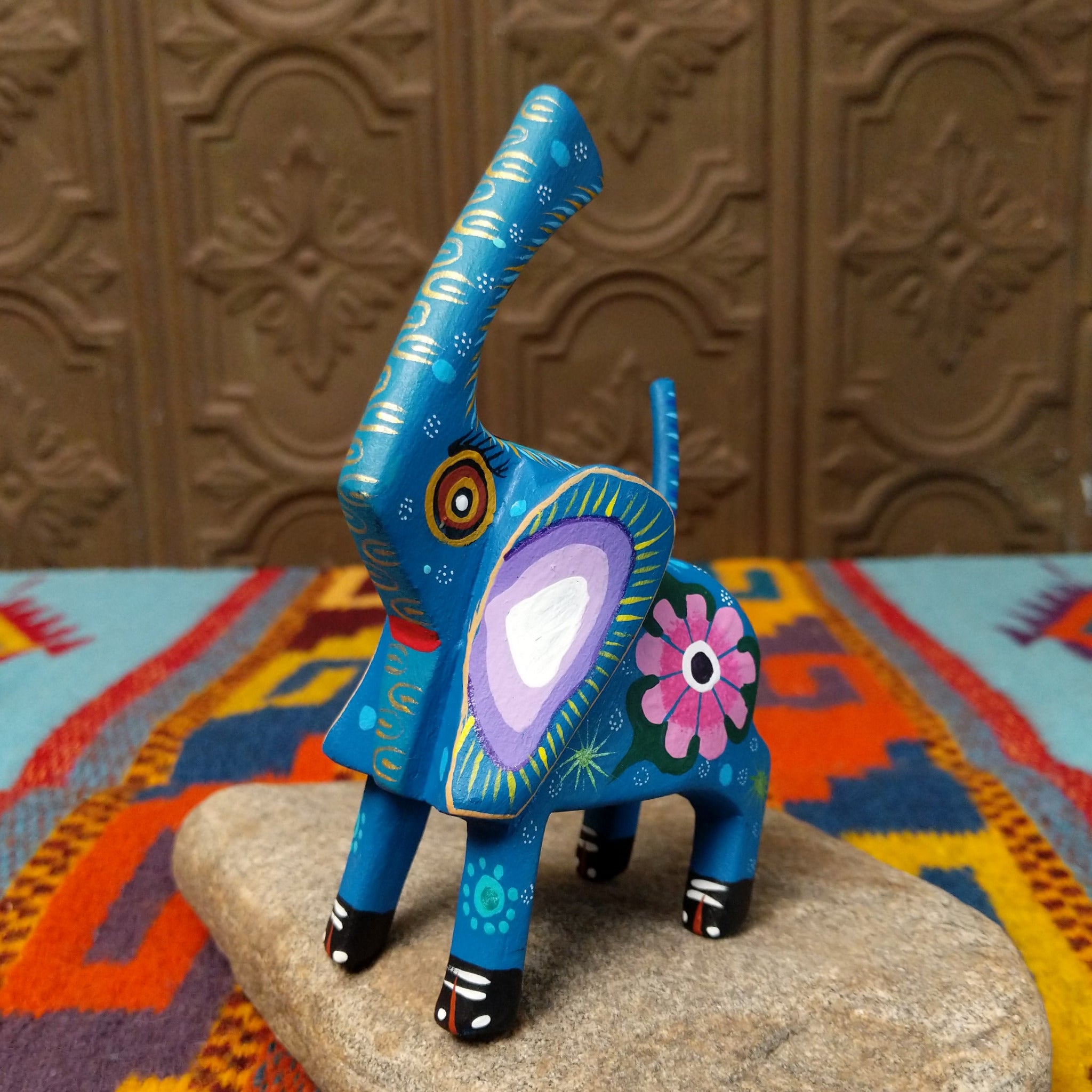 This blue Oaxacan elephant wood carving by Roberta Angeles features an uplifted trunk, lavender ear, pink flower, and colorful hand-painted decorative patterns.