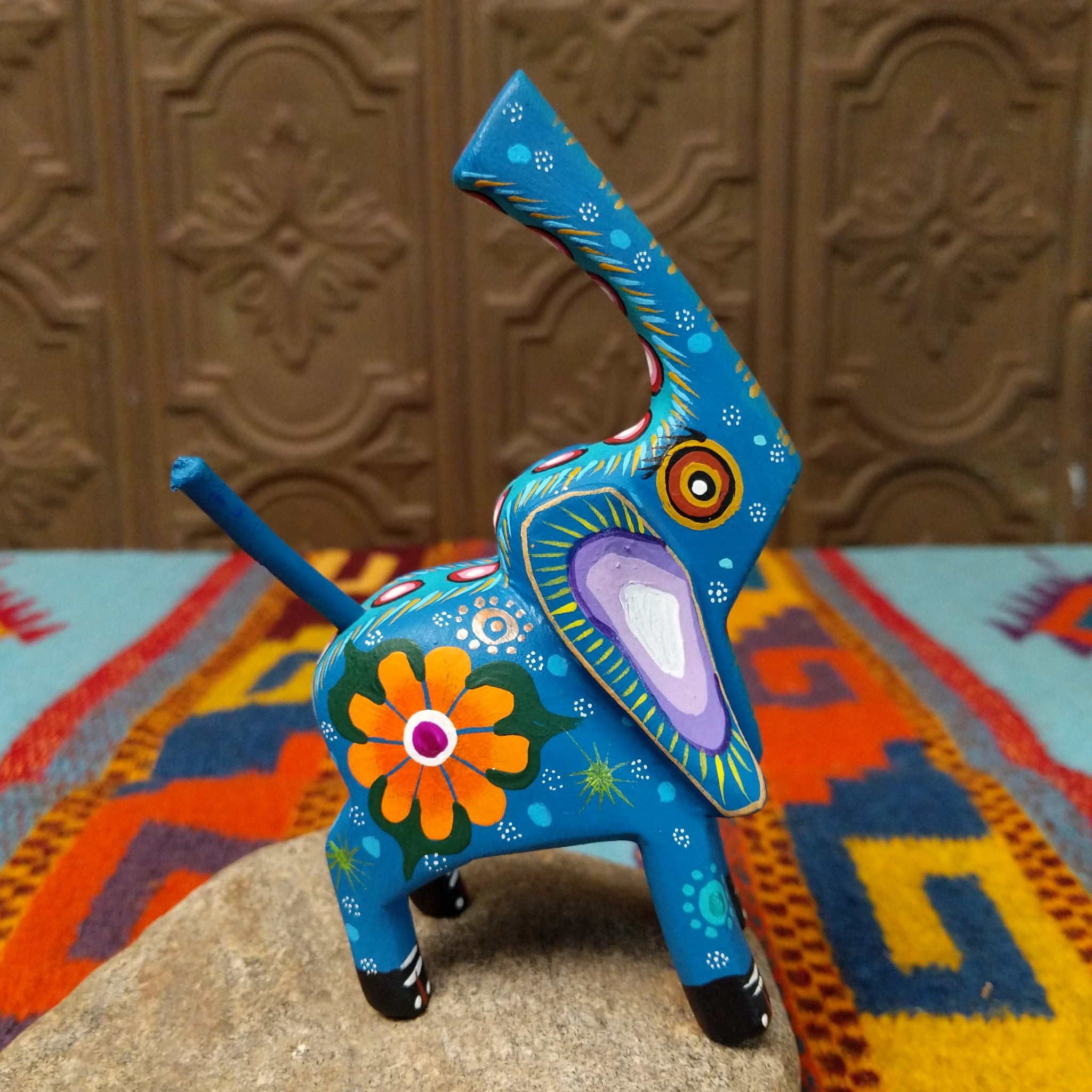 This blue Oaxacan elephant wood carving by Roberta Angeles features an uplifted trunk, lavender ear, pink flower, and colorful hand-painted decorative patterns; side view.