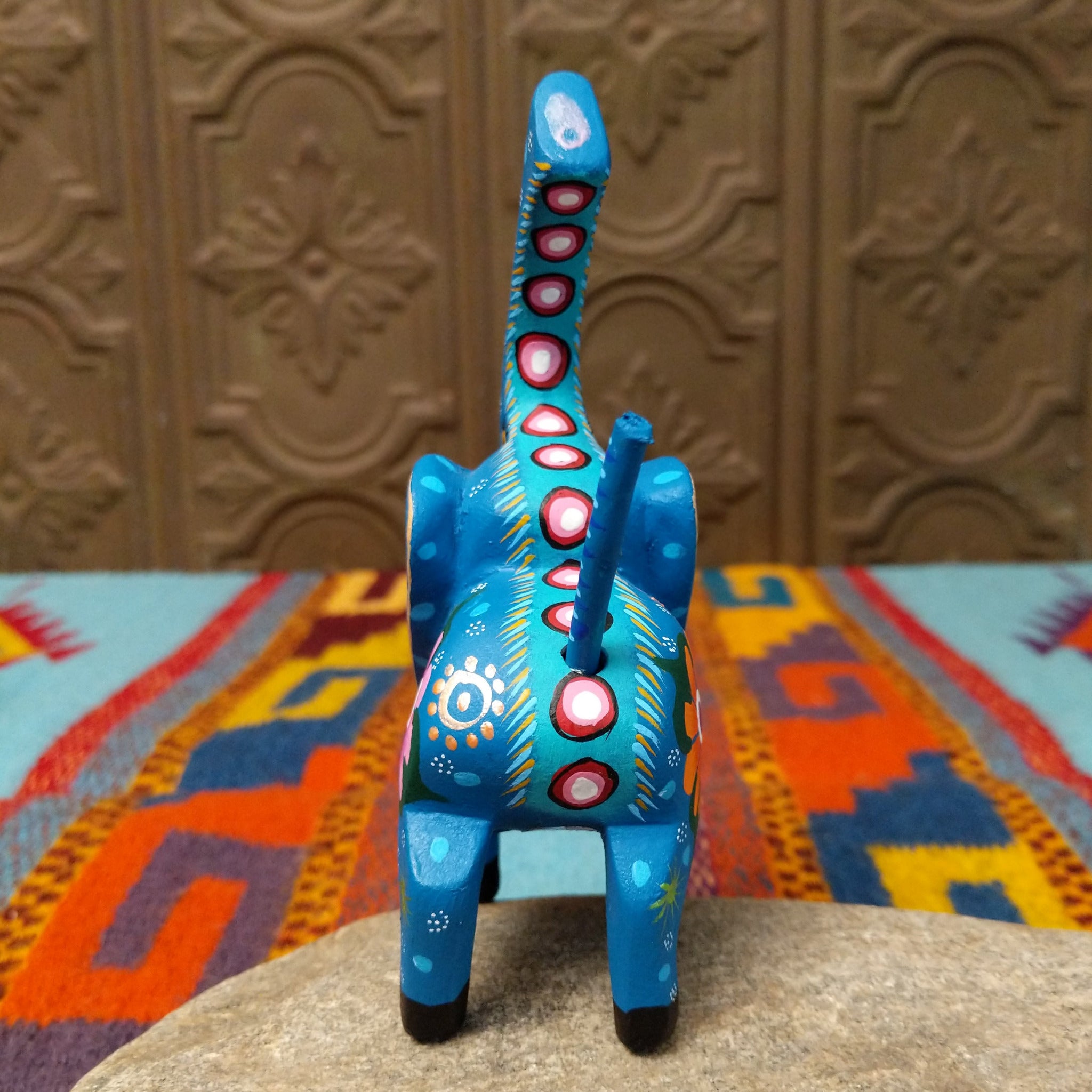 This blue Oaxacan elephant wood carving by Roberta Angeles features an uplifted trunk, lavender ear, pink flower, and colorful hand-painted decorative patterns; rear view.