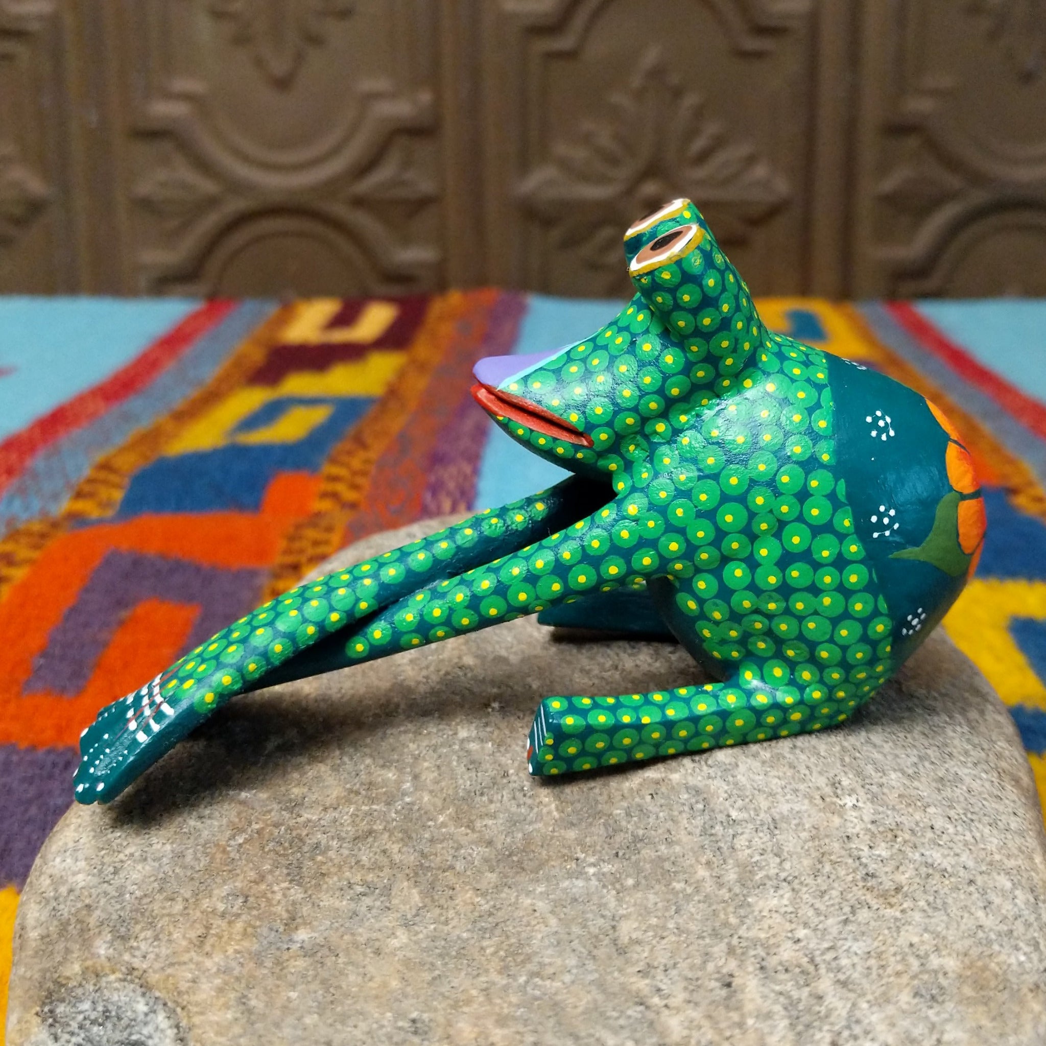 This green Oaxacan frog wood carving by Roberta Angeles features raised eyes, colorful dotted patterns, a red mouth, and long legs with oversized hand-painted webbed feet; side view.