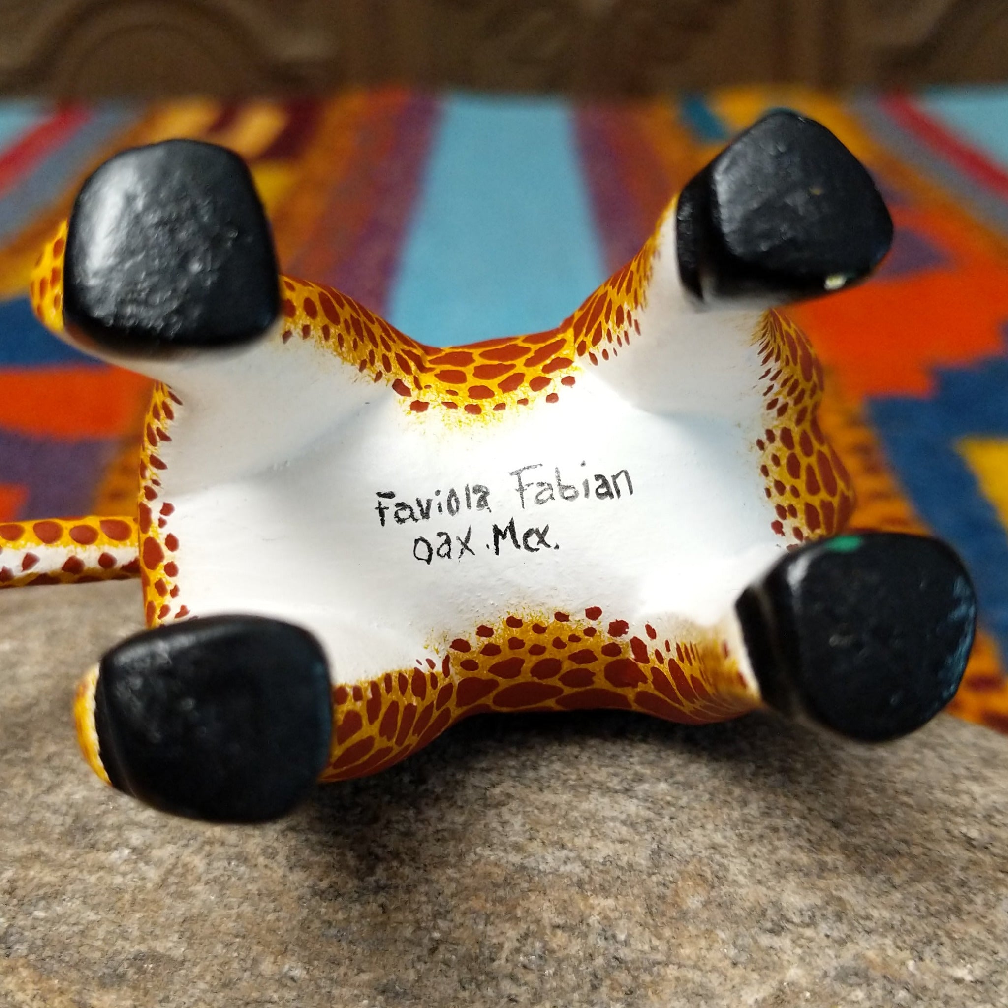 This Oaxacan giraffe wood carving by Faviola Fabian features a golden-yellow body with reddish-brown spots, a long neck, blue eyes, black-and-white horns, and black hooves; artisan name and location of production.