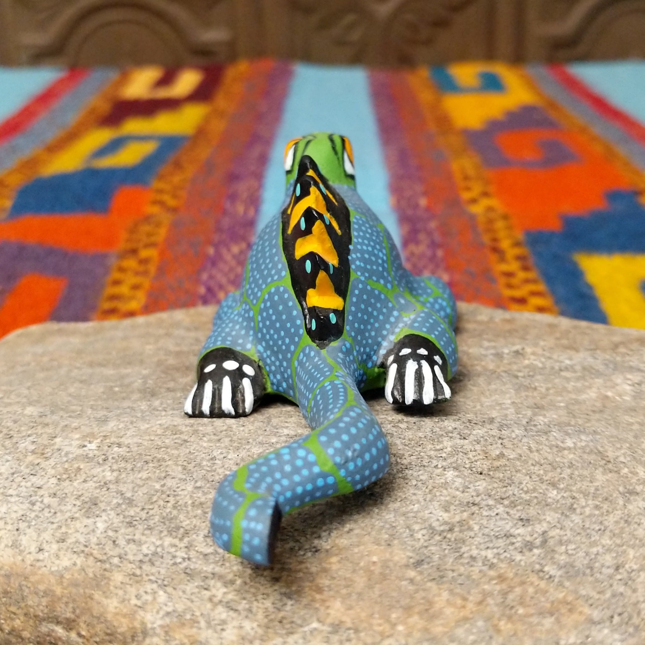 This colorful Oaxacan iguana wood carving features a green head, blue dotted body, golden eyes, black-and-yellow back spines, and a long patterned tail; rear view.
