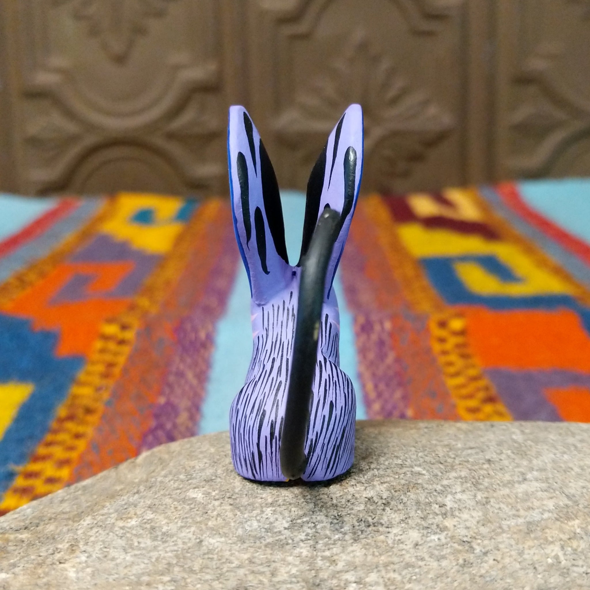 Oaxacan jerboa wood carving with oversized blue patterned ears, a lavender body, orange front legs, and long curved black tail.