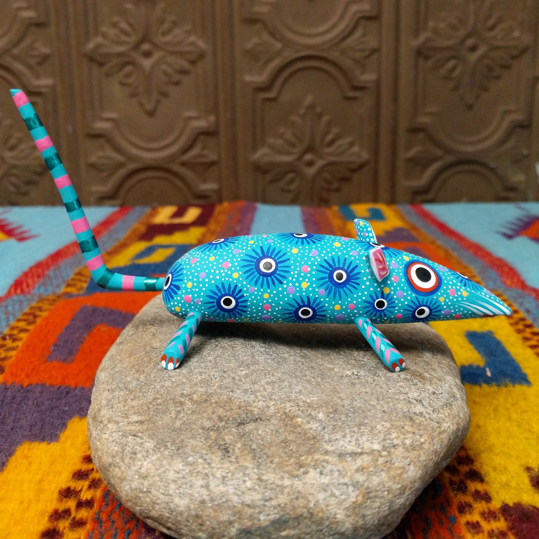 This turquoise Oaxacan mouse wood carving by Fabian Ramirez features colorful hand-painted patterns, pink-accented ears and legs, and a long striped tail; side view.