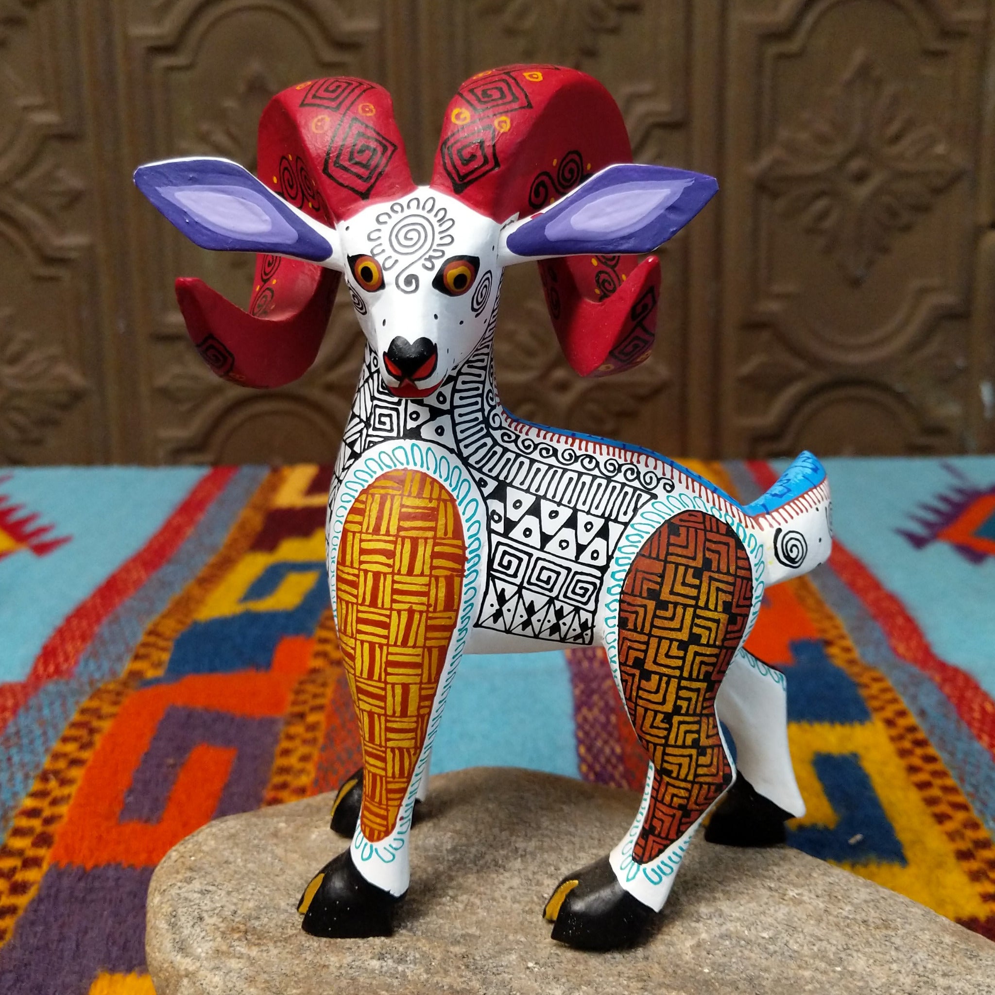 This Oaxacan ram wood carving by Abundia Munoz features large red curled horns, purple ears, a white body, and intricate black, orange, brown, and turquoise geometric patterns.