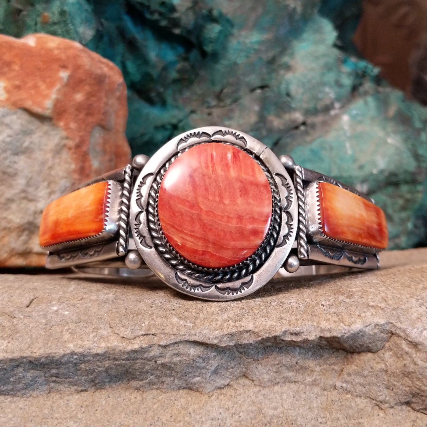 A sterling silver cuff features a central red spiny oyster shell stone and two orange spiny oyster shell pieces in notched bezels with twisted silver rope.