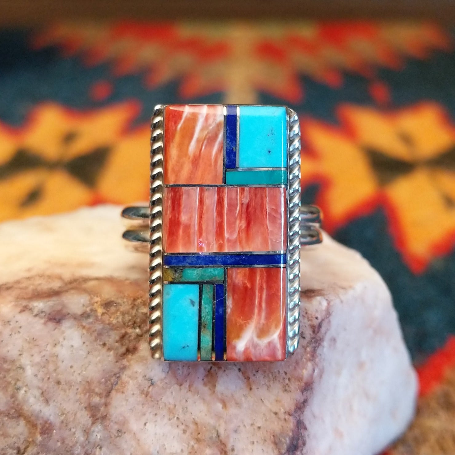 Size 10 ring by Wilbert Grey showcases an inlay design set in sterling silver. The rectangular face features a striking mosaic of vibrant stones, including rich red and orange spiney oyster shell, bright turquoise, and deep blue lapis. Front view