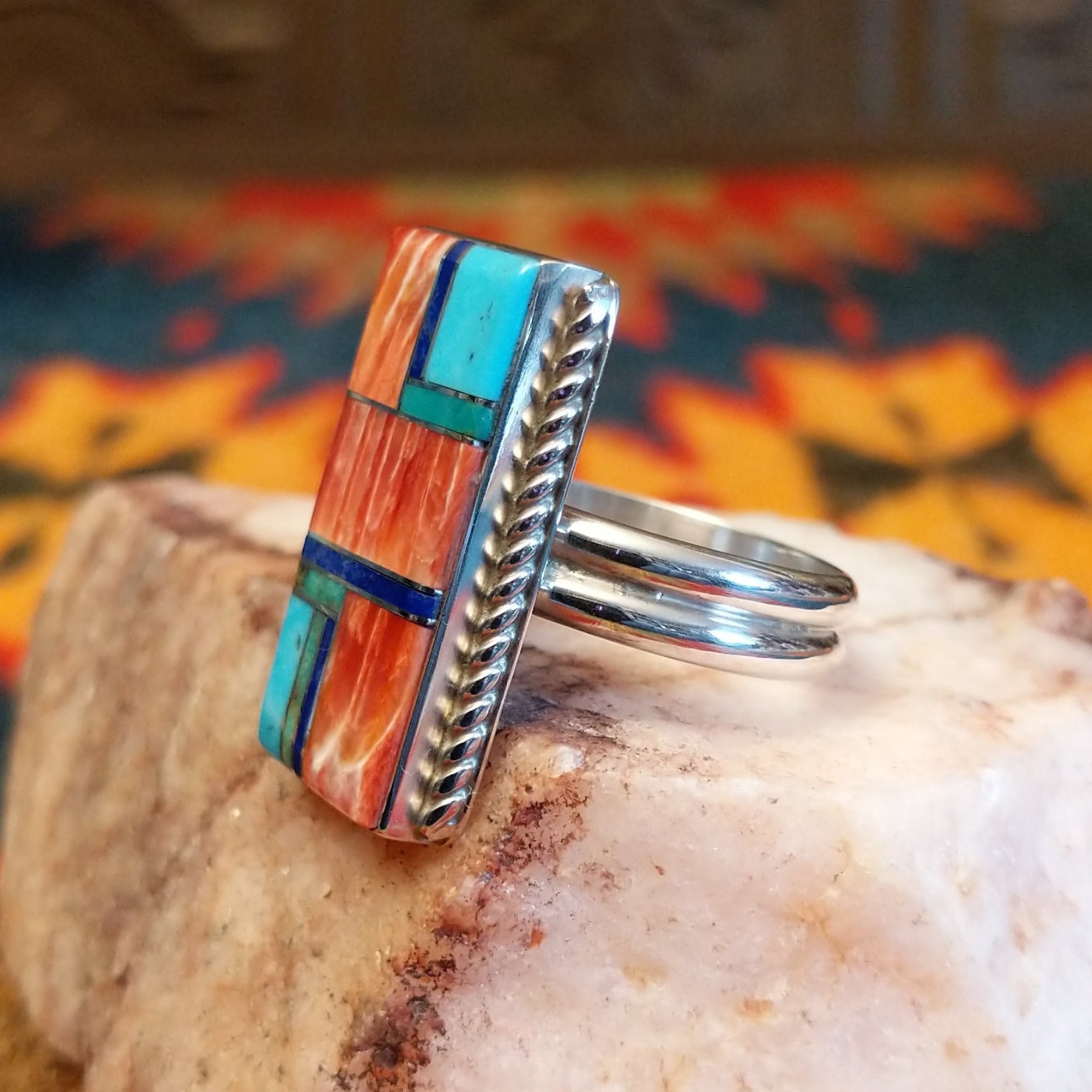 Size 10 ring by Wilbert Grey showcases an inlay design set in sterling silver. The rectangular face features a striking mosaic of vibrant stones, including rich red and orange spiney oyster shell, bright turquoise, and deep blue lapis. Side View