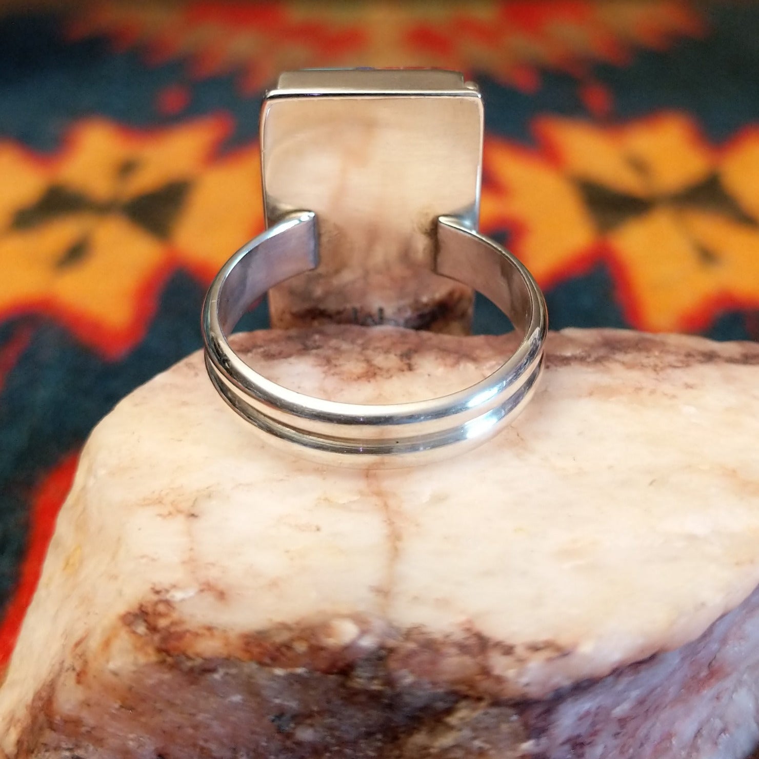 Size 10 ring by Wilbert Grey showcases an inlay design set in sterling silver. The rectangular face features a striking mosaic of vibrant stones, including rich red and orange spiney oyster shell, bright turquoise, and deep blue lapis. Underside of ring view