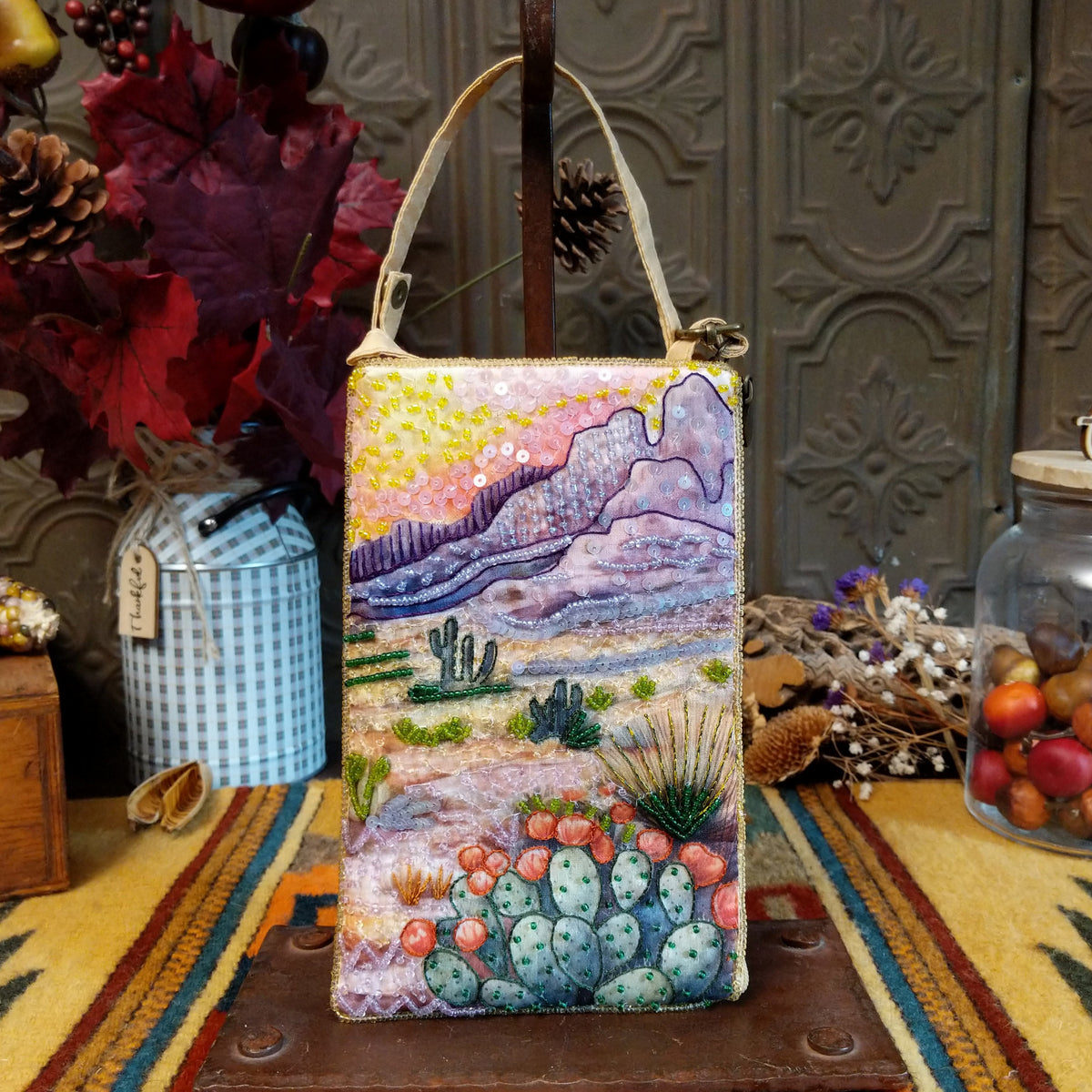 Prickly Pear Club Bag by Bamboo Trading featuring a colorful beaded Southwestern desert landscape with purple mountains, pink and yellow sunset sky, prickly pear cacti with coral-colored blossoms, desert plants, sparkling sequins, and a cream-colored shoulder strap.