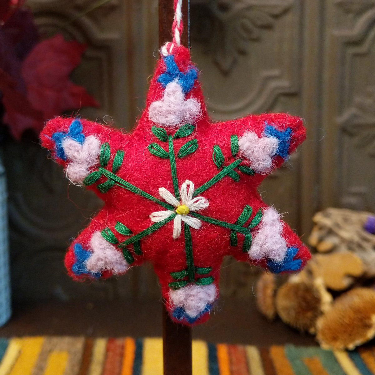 This charming Red Star Wool Ornament features a vibrant red six-pointed star with embroidered green branches, a white flower with a yellow center, soft pink floral accents, blue details, and a red and white twisted hanging cord.