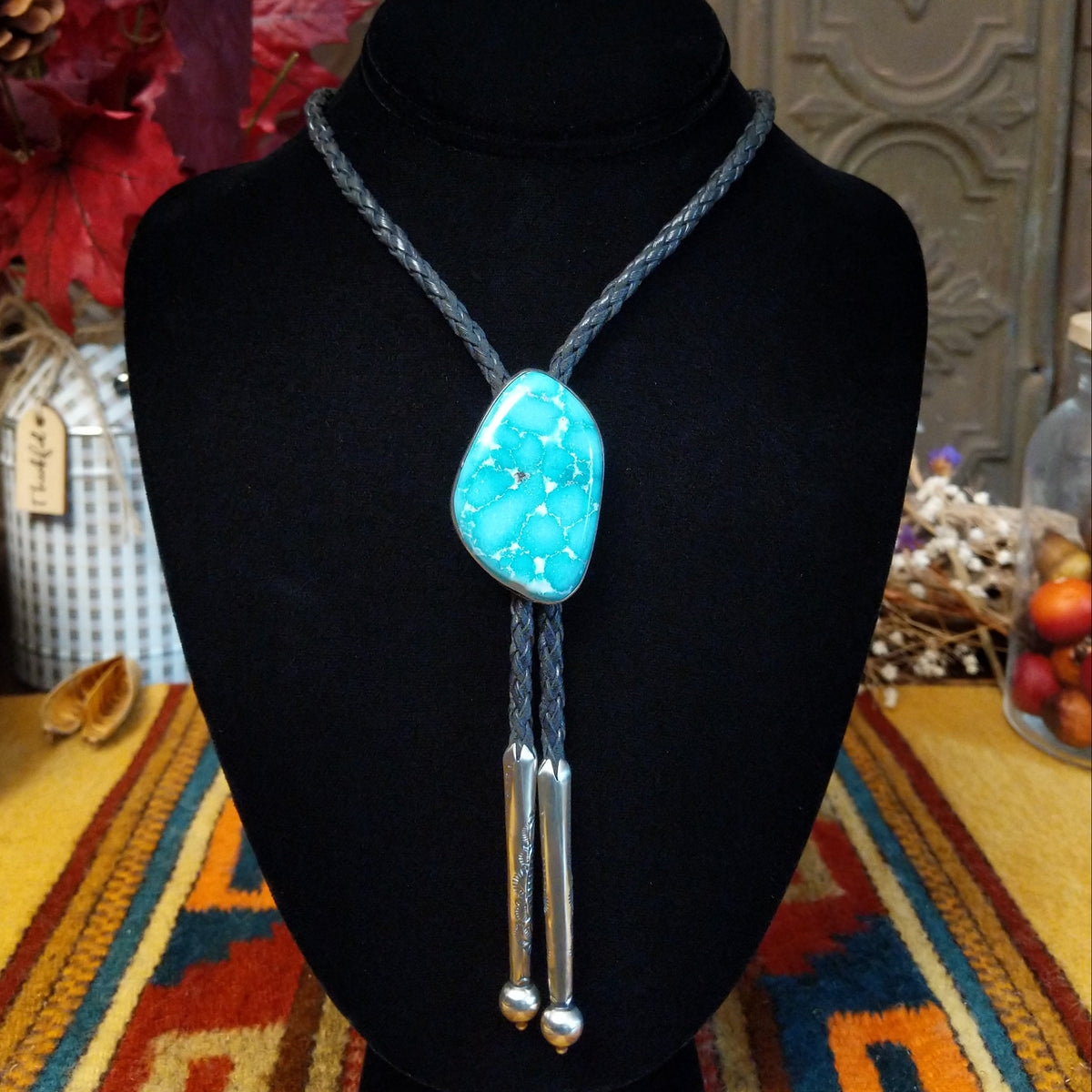 A turquoise stone bolo tie necklace with a black braided cord and Sterling Silver dangling tips, displayed on a black velvet mannequin bust.