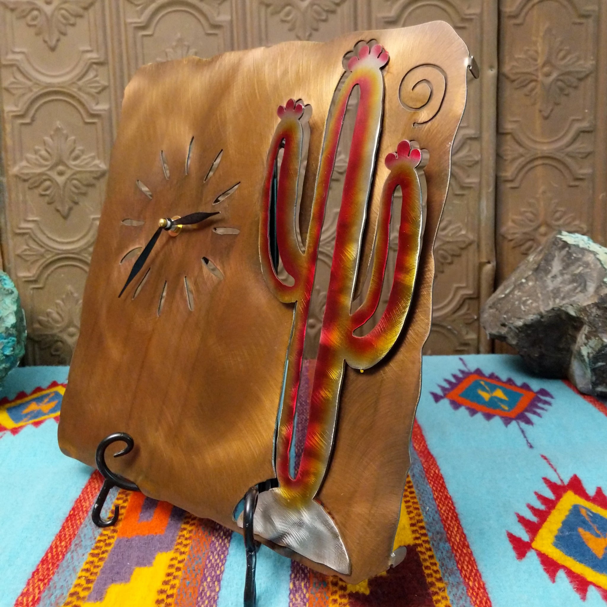 Handcrafted Crooks Design metal clock featuring a colorful red, yellow, and cream saguaro cactus on a brushed warm-gold background with cutout hour markers and black clock hands; side view.