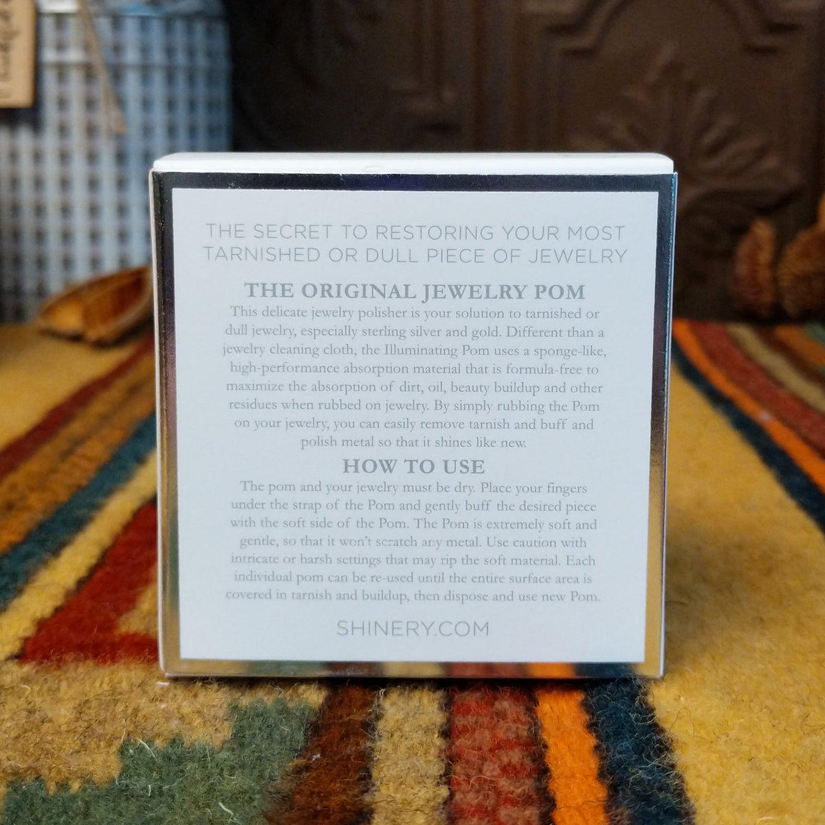 A small square product box standing upright on a patterned rug, with printed instructions on the front. The background shows a decorative interior with a carved wooden door, a blue-and-white vase with red leaves, and dried floral decor.