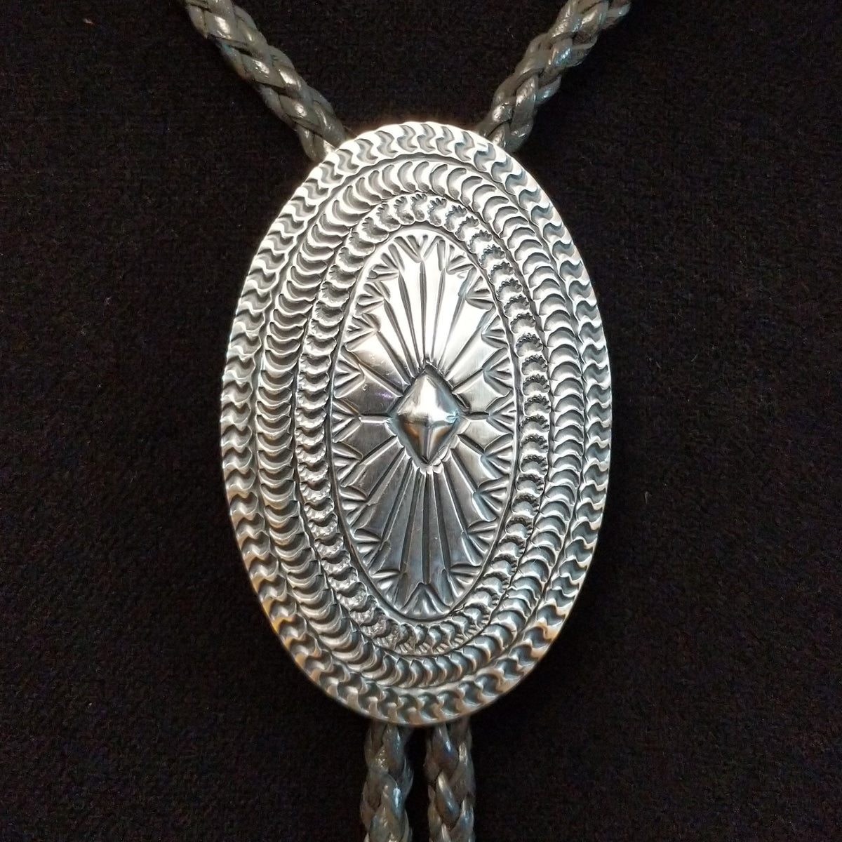 A silver-toned oval bolo tie pendant with intricate engraved patterns and a braided cord, displayed against a black background.