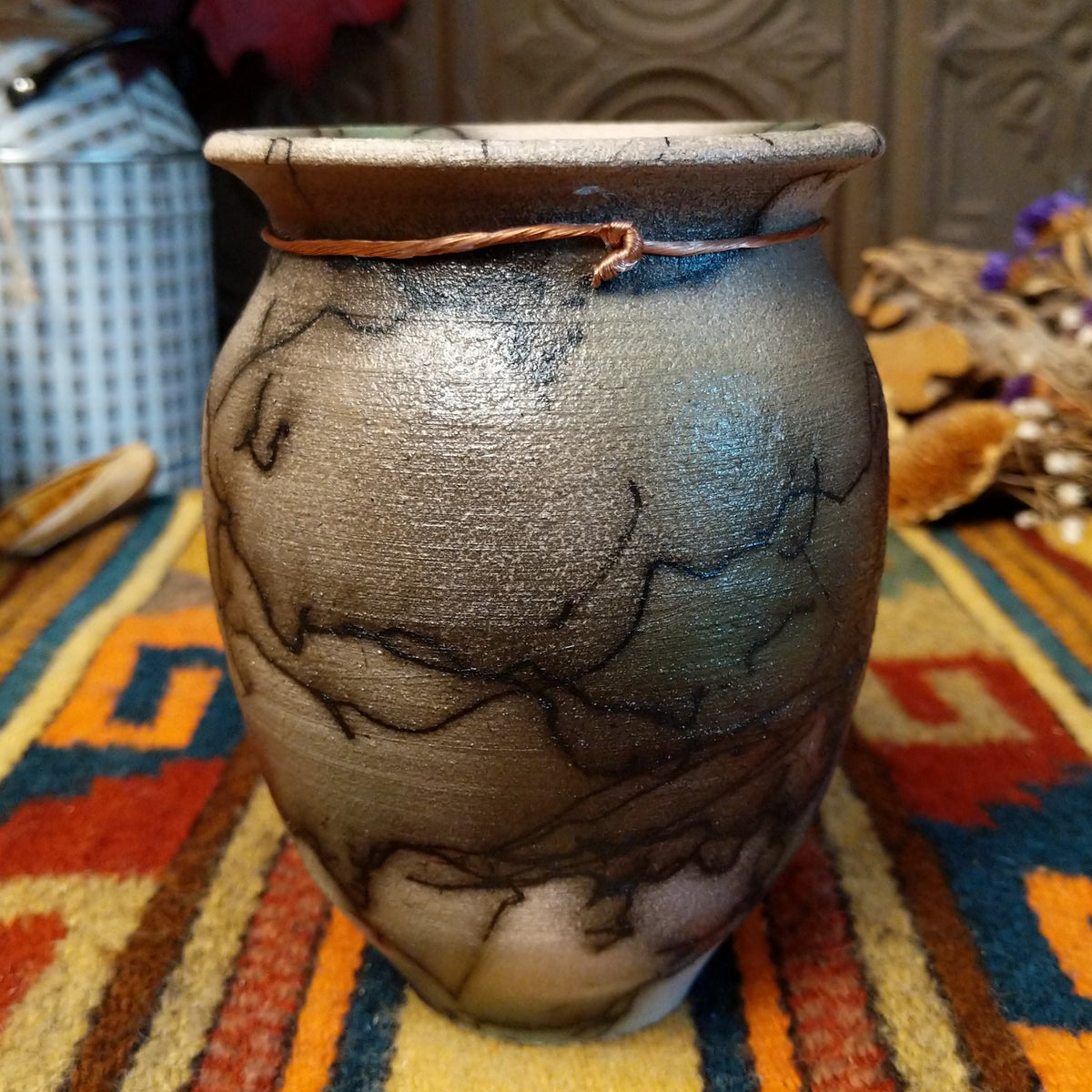 This elegant Small Mottled Horsehair Pottery by Roger Calhoun features a rounded ceramic vase with earthy tones, subtle green accents, black horsehair patterns, a flared rim, and decorative copper wire with a green patterned bead.