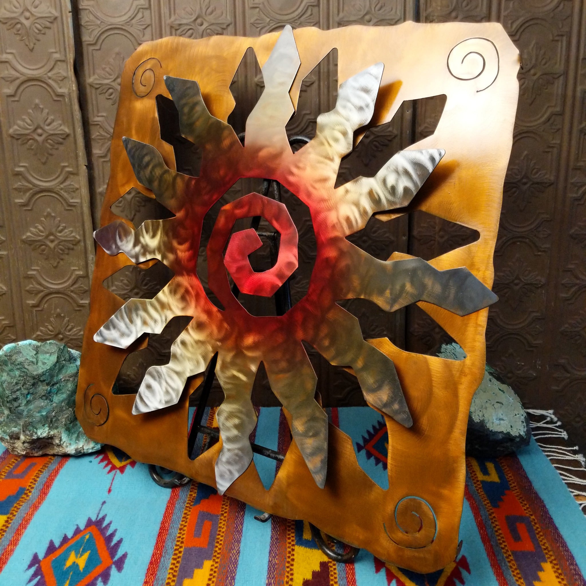 This Crooks Design metal wall art features a 12-point sunburst with angular silver and gold rays, a deep red spiral center, and a warm golden-brown frame with spiral accents; side view.