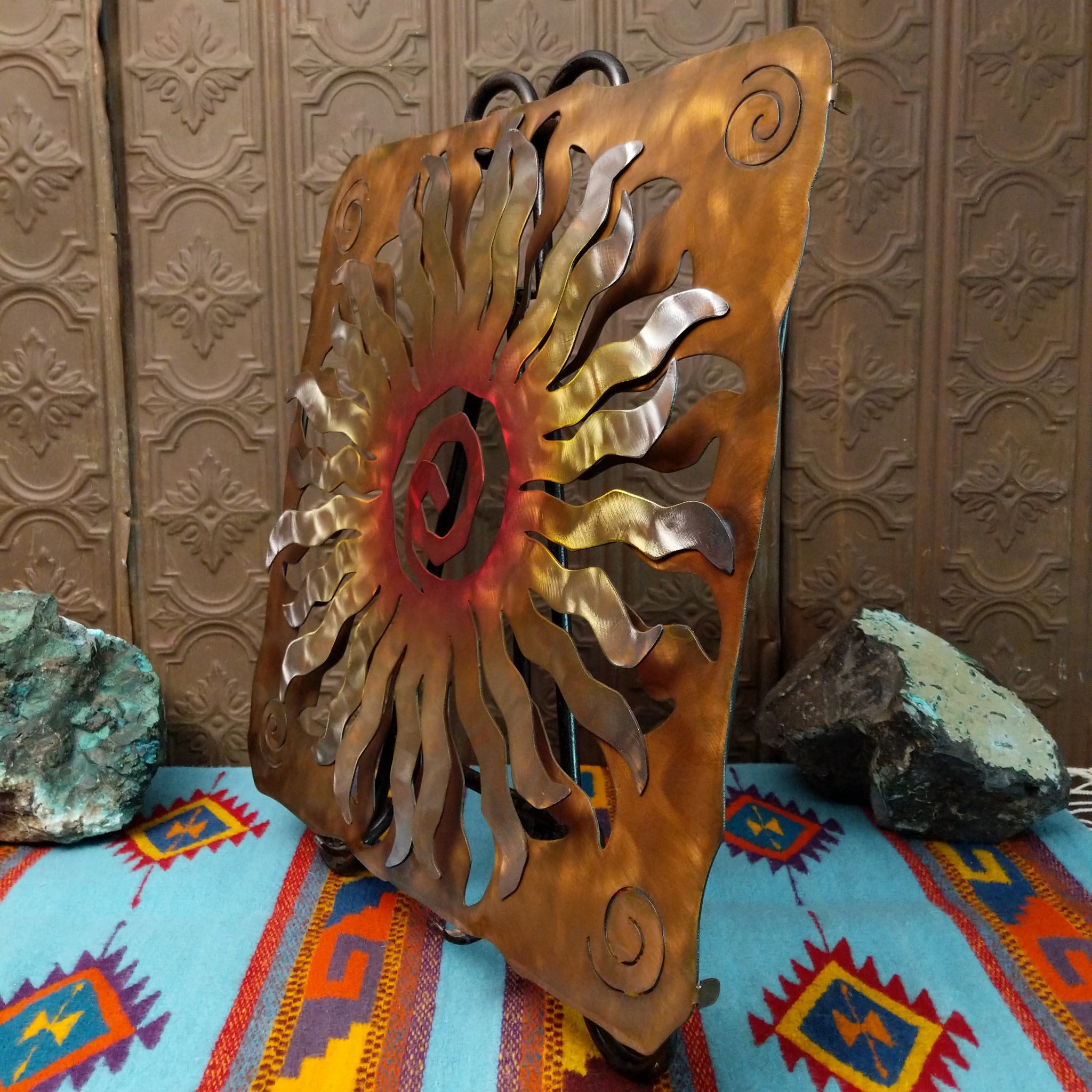 This Crooks Design metal wall art features a 24-point sunburst with flowing silver and gold rays, a red and orange spiral center, and a warm golden-brown frame with spiral accents; side view.