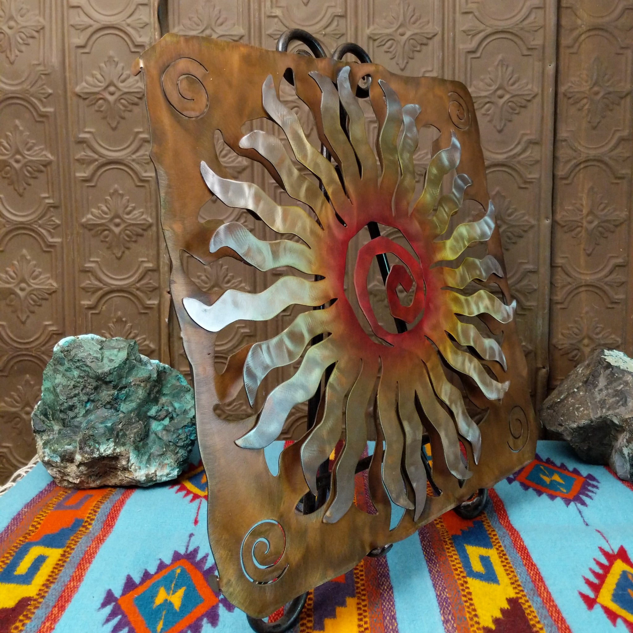 This Crooks Design metal wall art features a 24-point sunburst with flowing silver and gold rays, a red and orange spiral center, and a warm golden-brown frame with spiral accents; side view.