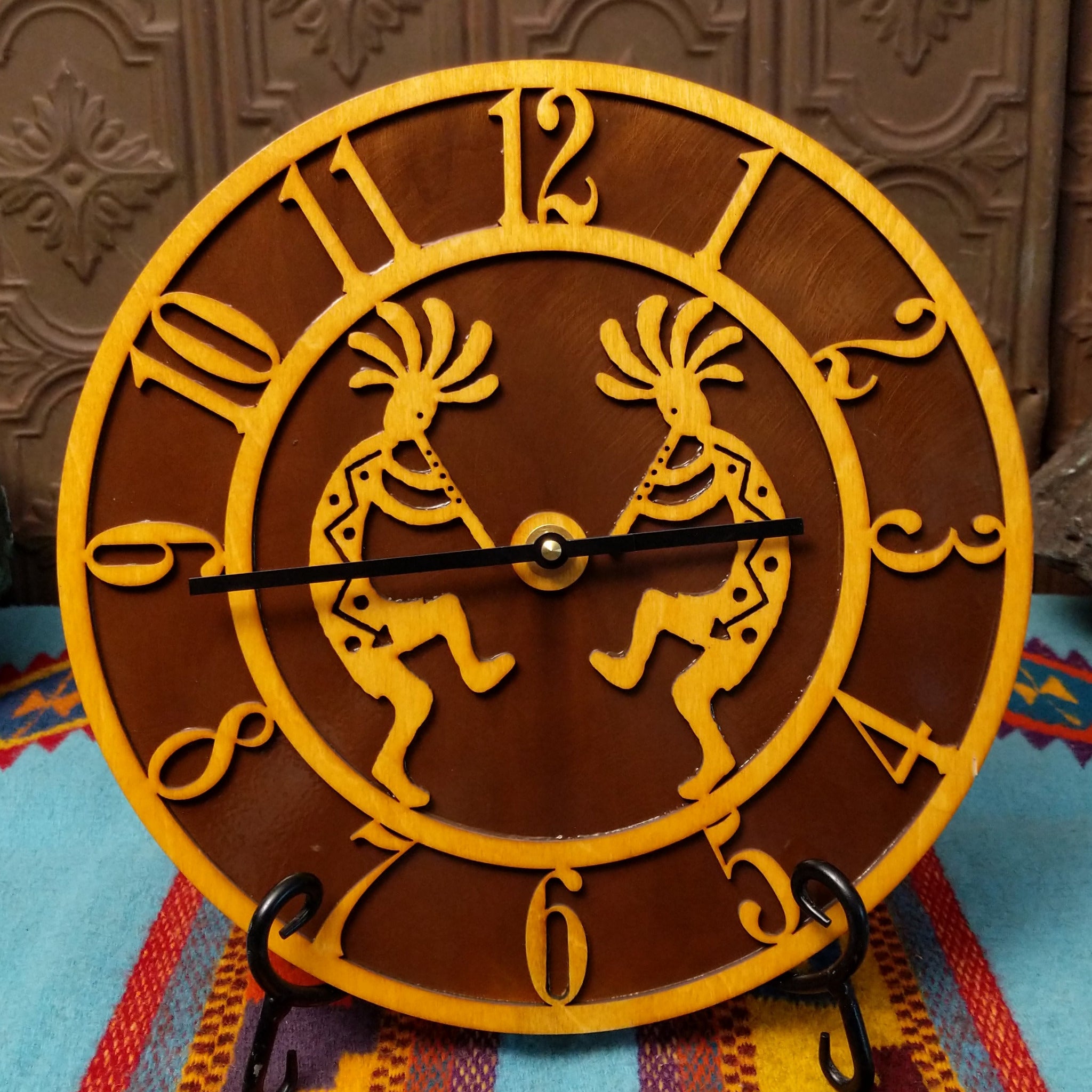 This Crooks Design round Tableau Clock features a pair of golden-toned Kokopelli figures, stylized openwork numerals, black clock hands, and a rich brown background.