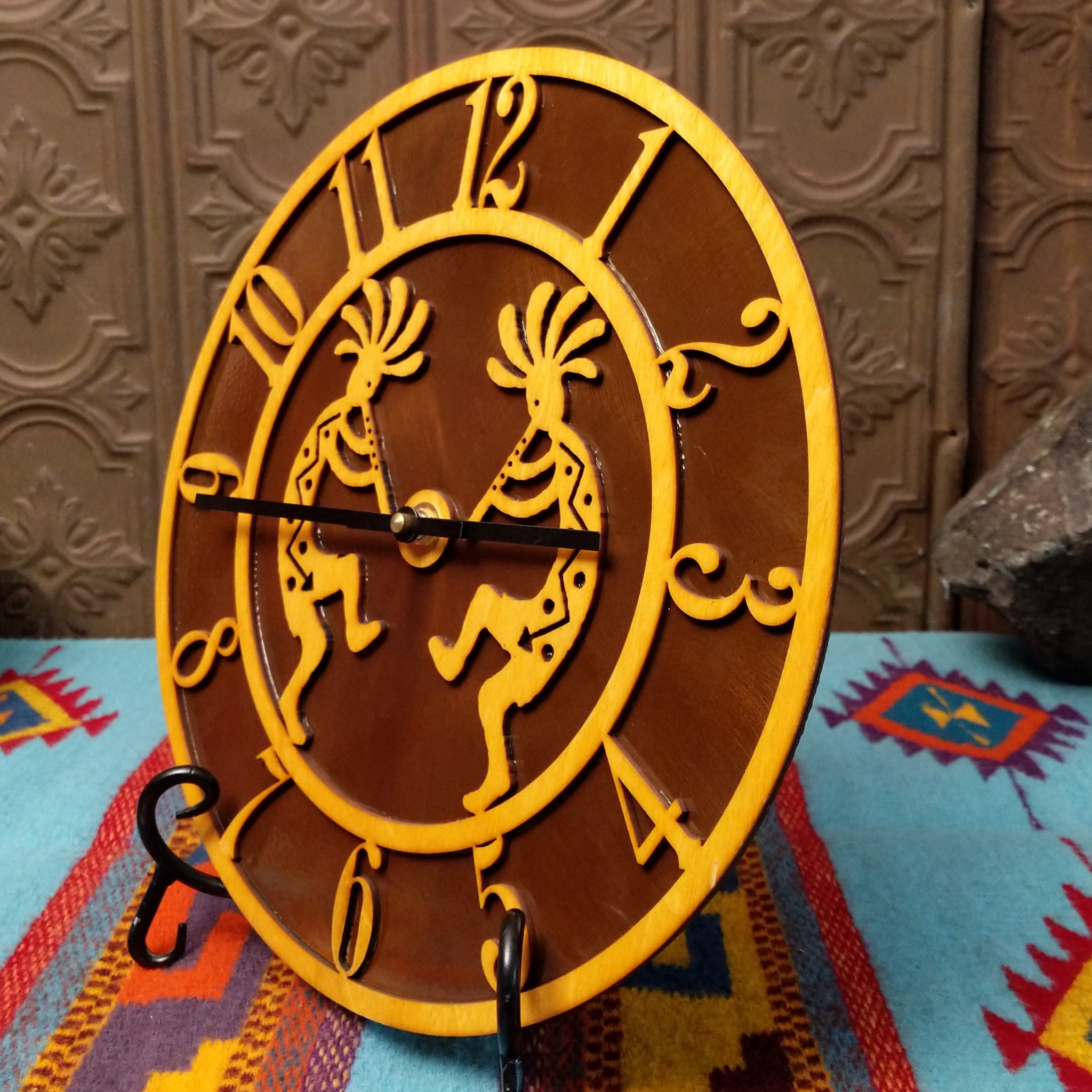This Crooks Design round Tableau Clock features a pair of golden-toned Kokopelli figures, stylized openwork numerals, black clock hands, and a rich brown background; side view.