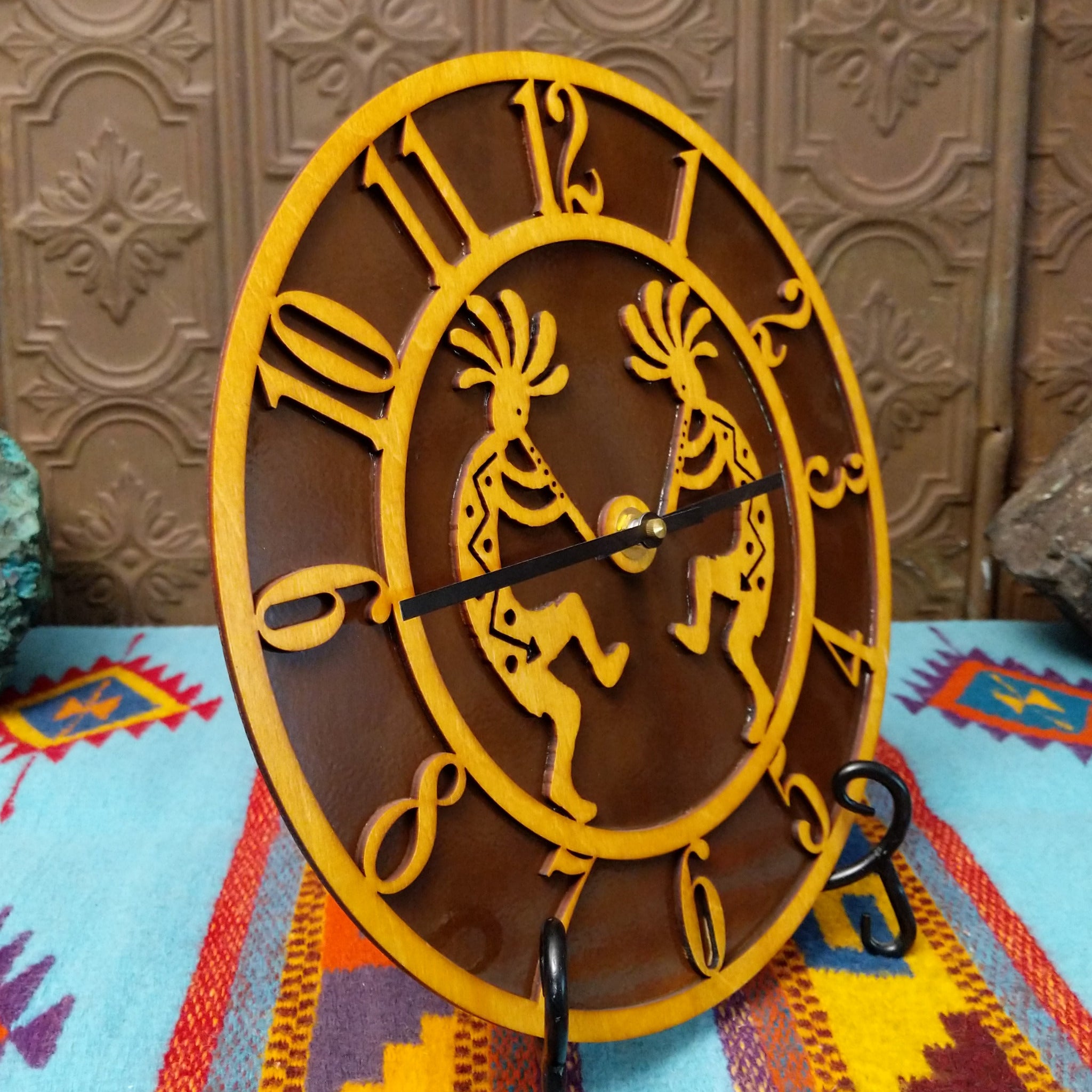 This Crooks Design round Tableau Clock features a pair of golden-toned Kokopelli figures, stylized openwork numerals, black clock hands, and a rich brown background; side view.