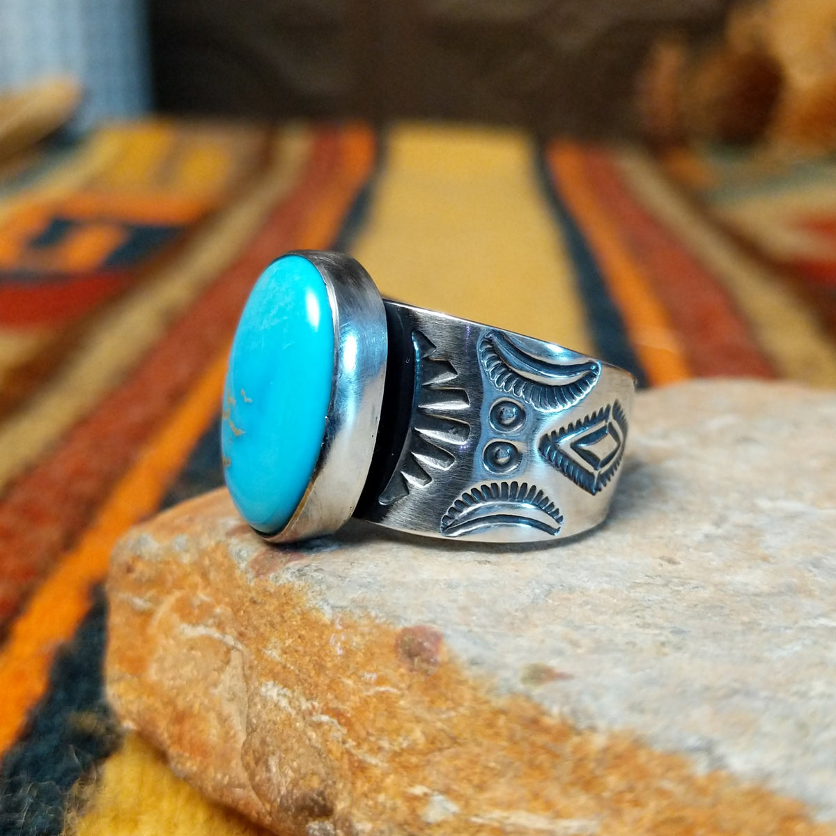 Turquoise Ring by Lowell Draper & Bernyse Chavez featuring a large oval turquoise stone with natural matrix, a polished silver bezel, and a wide silver band decorated with stamped sunburst patterns and geometric detailing; side view.