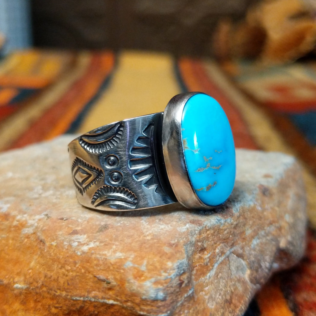 Turquoise Ring by Lowell Draper & Bernyse Chavez featuring a large oval turquoise stone with natural matrix, a polished silver bezel, and a wide silver band decorated with stamped sunburst patterns and geometric detailing; side view.