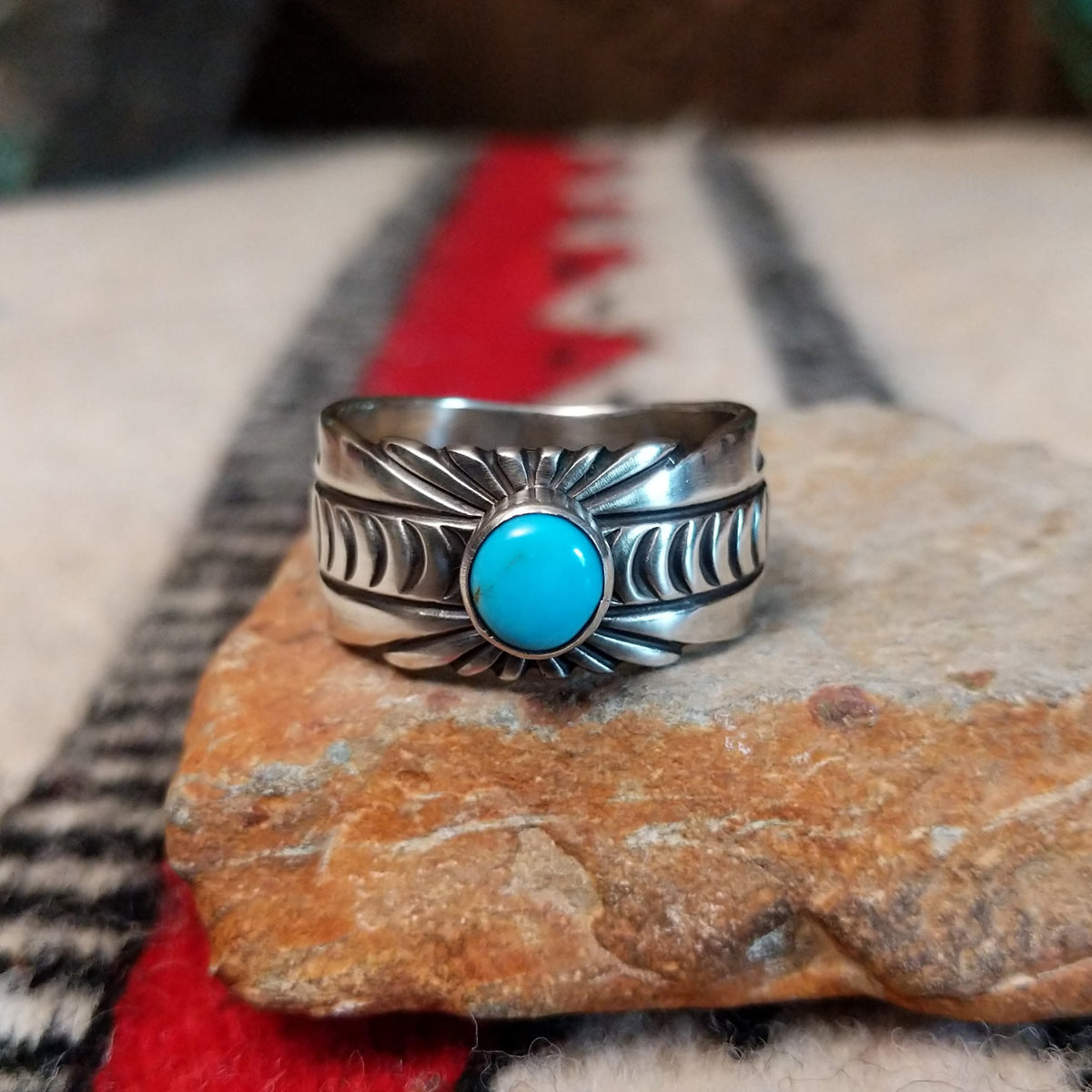 Turquoise ring by Raymond Coriz featuring a round turquoise stone centered in a wide silver band with detailed stamped, radiating, and curved designs.