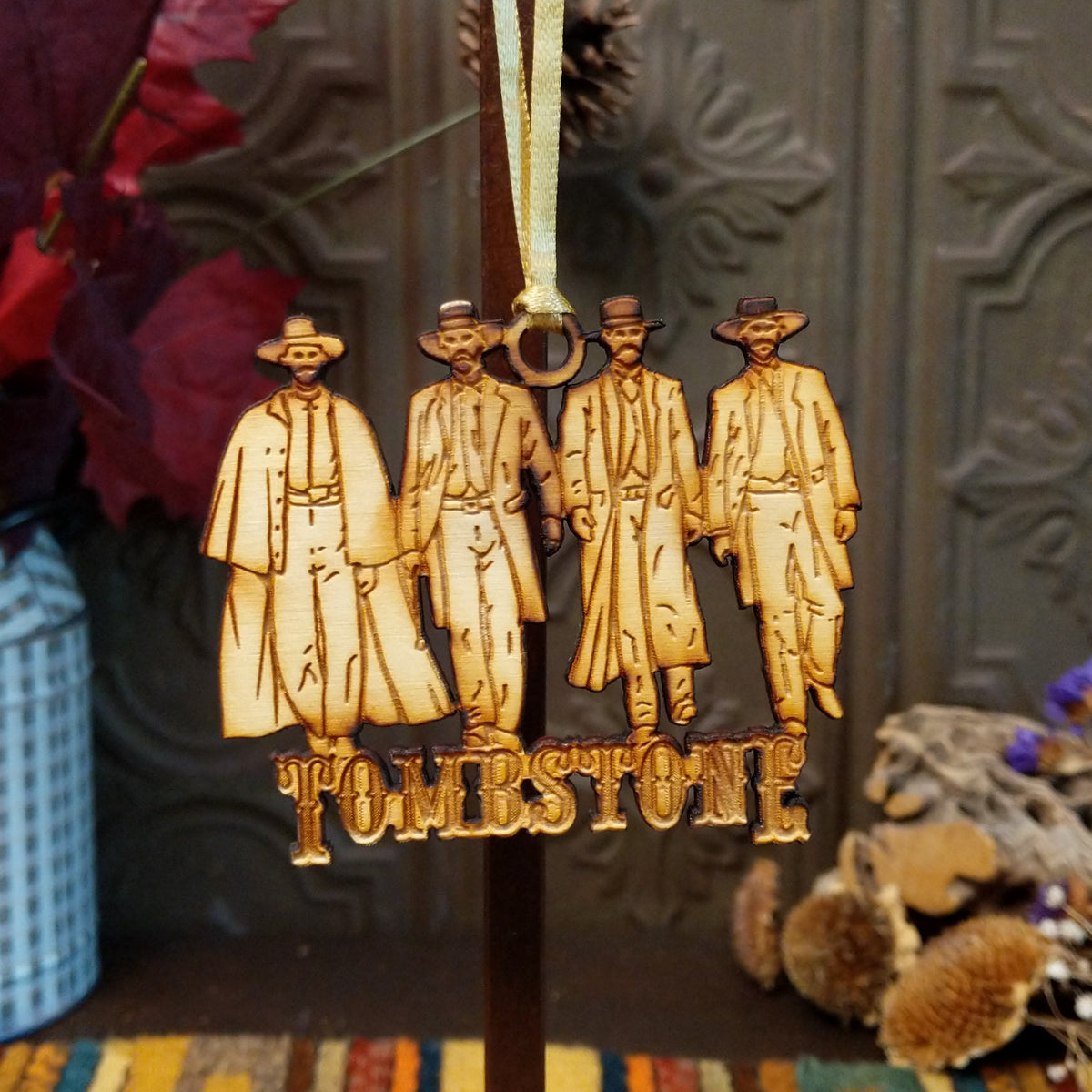 A wooden hanging ornament featuring four engraved figures in cowboy hats and long coats, with the word "TOMBSTONE" below, suspended by a gold ribbon against a dark decorative background.