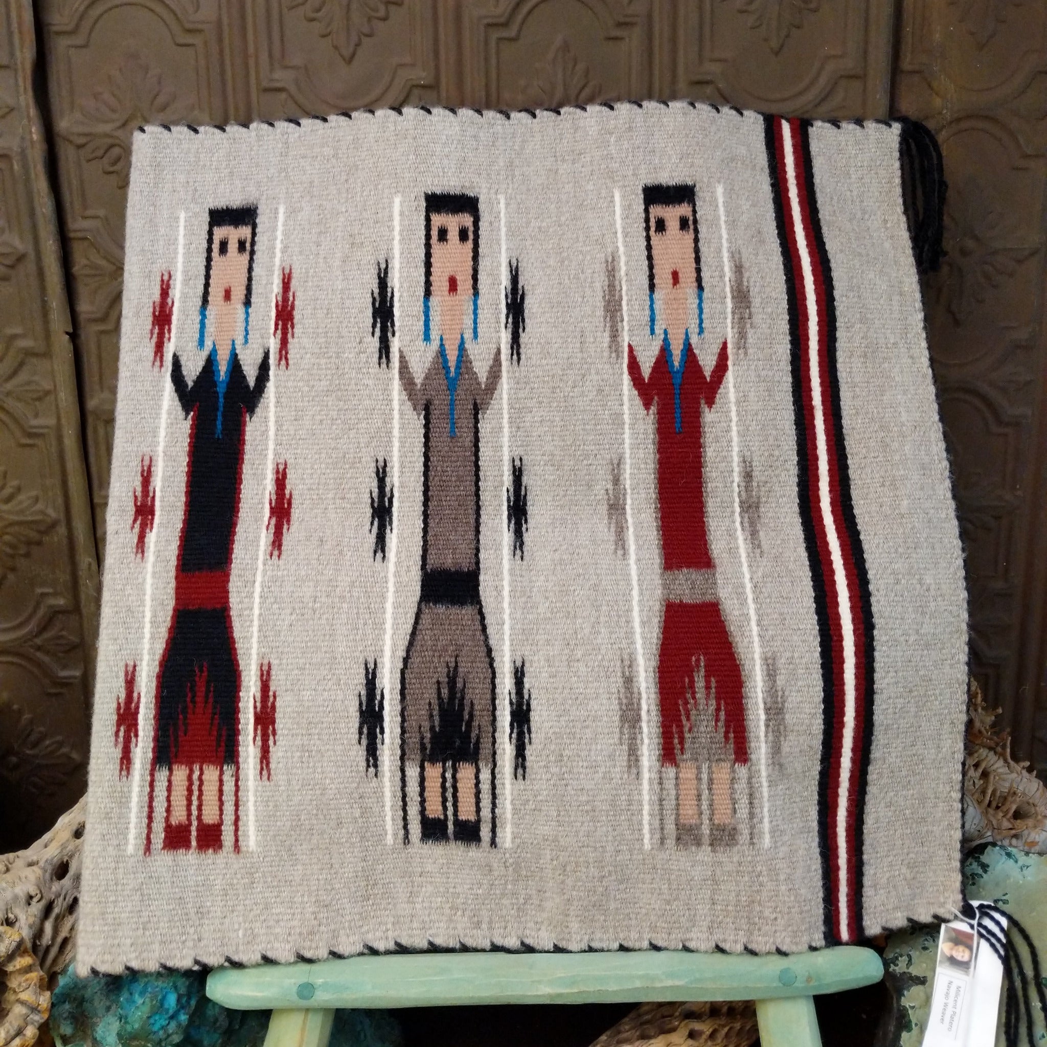 Yei Navajo rug by Milicent Platero featuring three traditional Yei figures on a natural gray background with geometric accents and a striped side border.