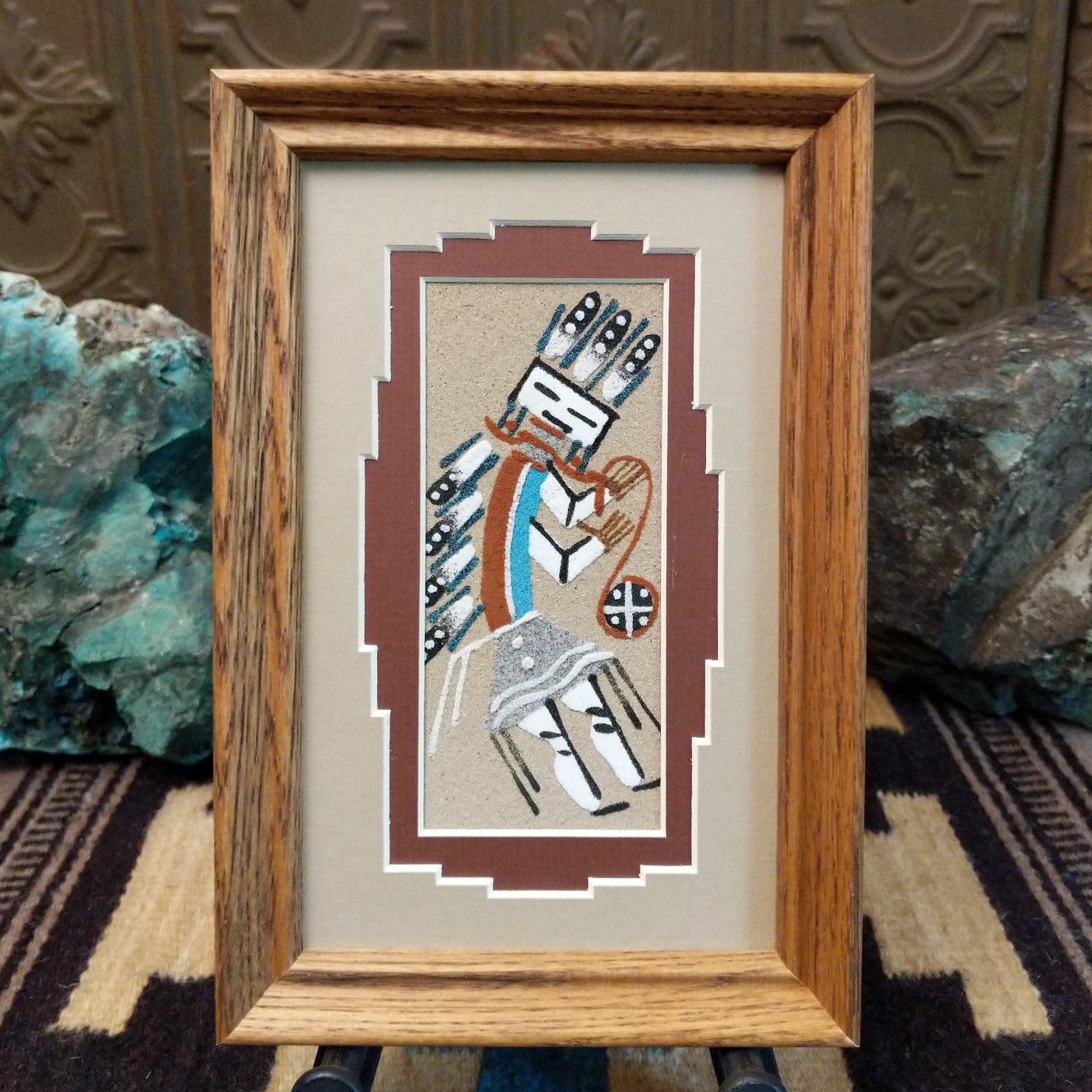 Framed Navajo sand painting of a side-facing Yei figure with turquoise accents and feather details.