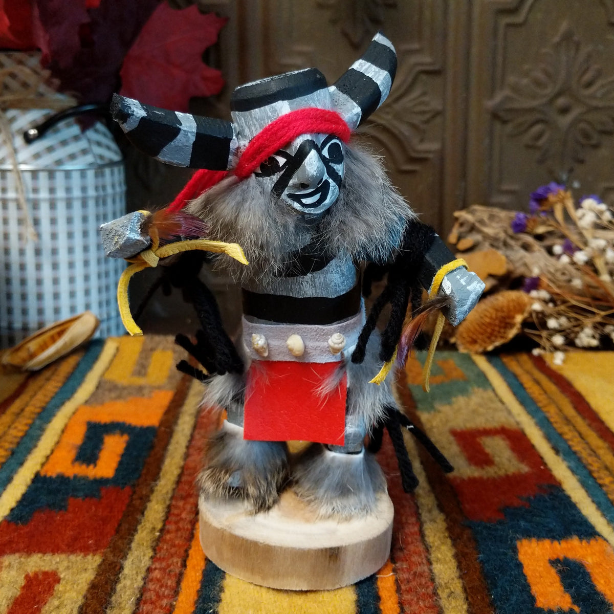 Navajo Clown Kachina doll with black-and-silver striped horns, smiling painted face, gray fur, striped body, and red fabric apron.