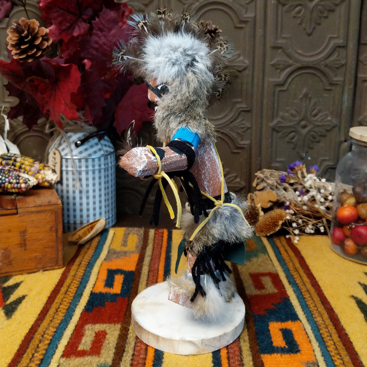 Side view of the Navajo Protection Dancer Kachina doll with gray fur headdress, black painted facial markings, blue sleeves, feather accents, and raised foot.