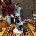 Side view of the Navajo Sunface Kachina doll with turquoise-colored face, feather headdress, outstretched arms, and blue fabric clothing.