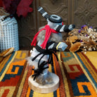 Side view of the Navajo Clown Kachina doll with black-and-silver striped horns, smiling painted face, gray fur, striped body, and red fabric apron.