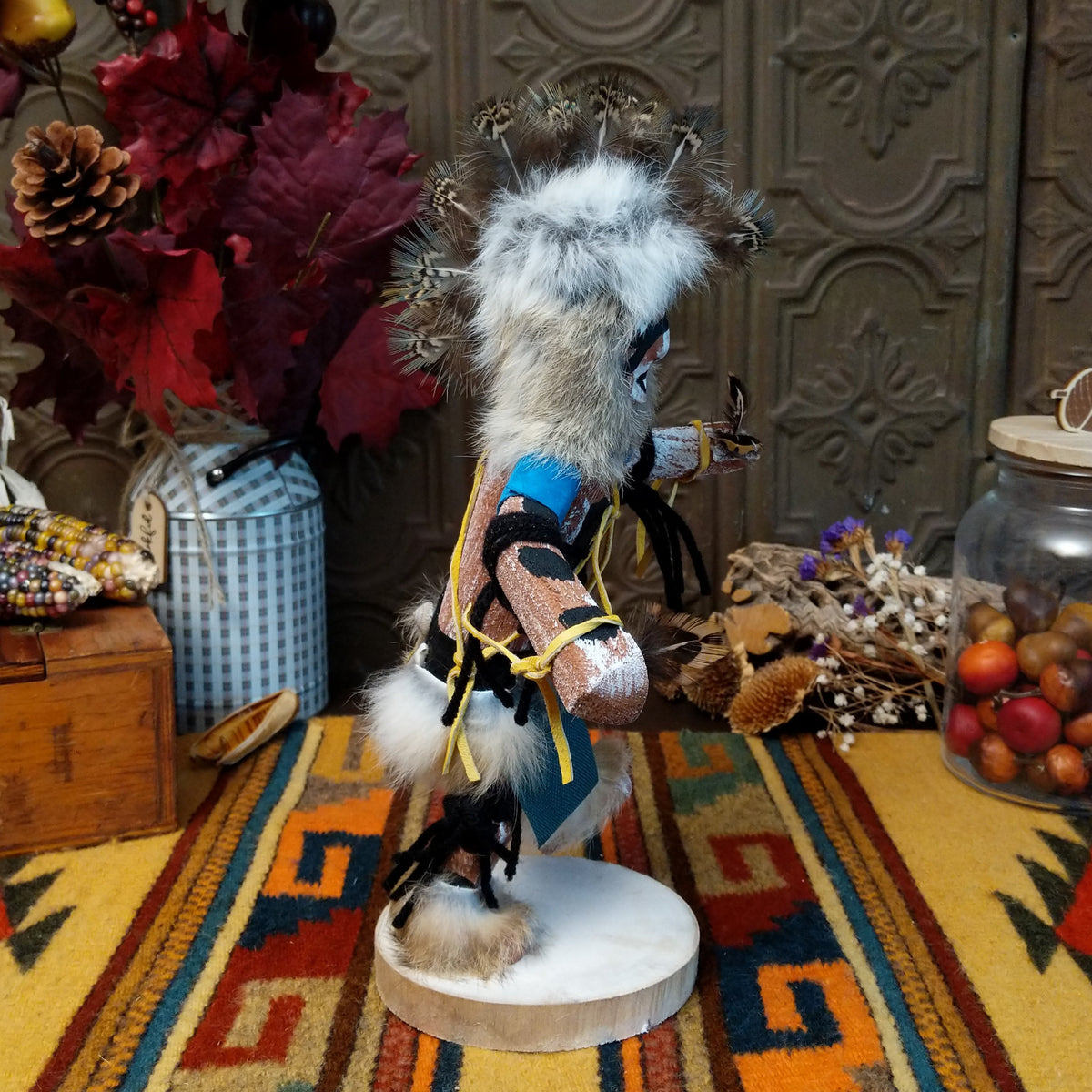 Side view of the Navajo Protection Dancer Kachina doll with gray fur headdress, black painted facial markings, blue sleeves, feather accents, and raised foot.