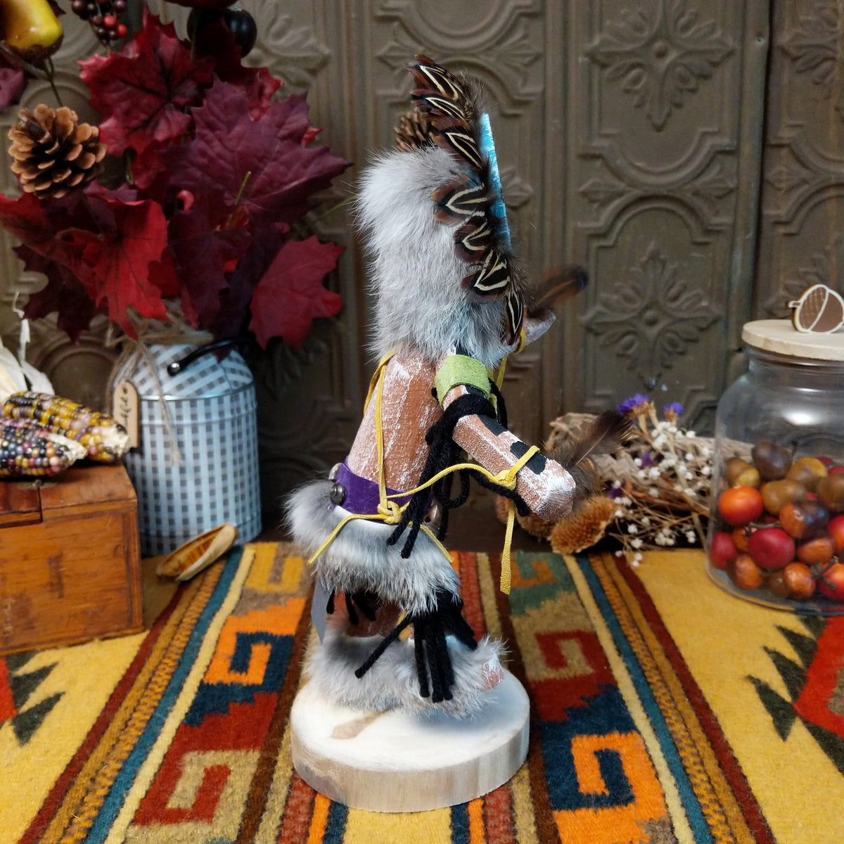 Side view of the Navajo Sunface Kachina doll with turquoise-colored face, feather headdress, outstretched arms, and blue fabric clothing.