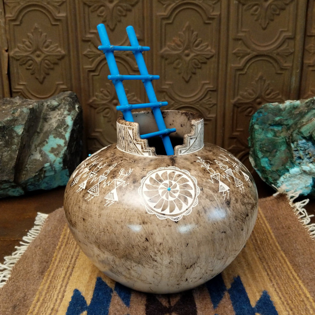 White clay pottery with fired horsehair and intricate etched designs of friendship dancers on the surface, stepped lip, and blue, wooden ladder.