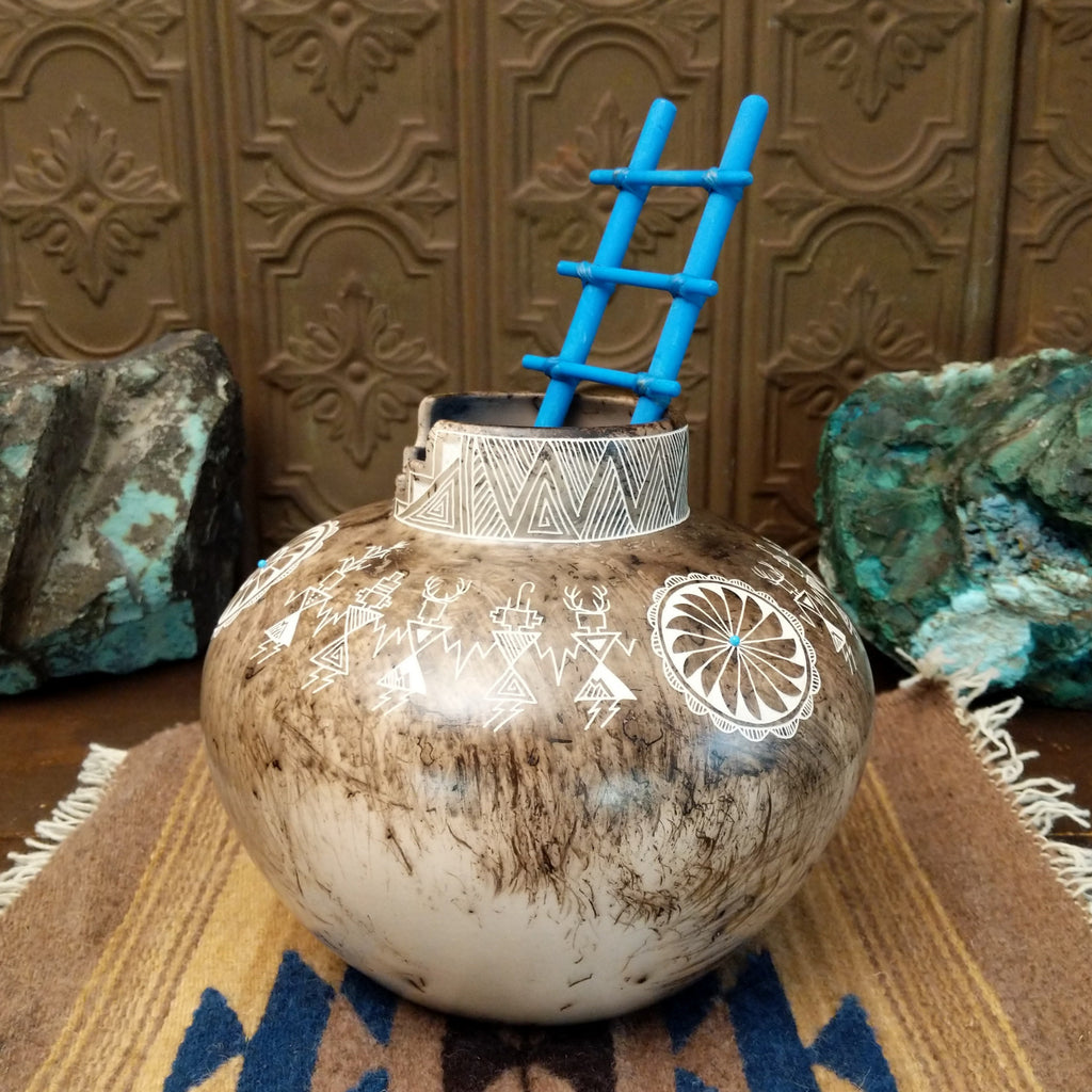 White clay pottery with fired horsehair and intricate etched designs of friendship dancers on the surface, stepped lip, and blue, wooden ladder.