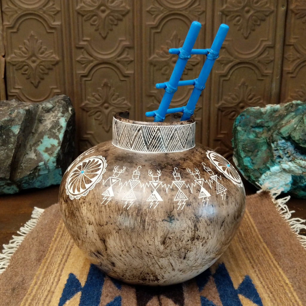 White clay pottery with fired horsehair and intricate etched designs of friendship dancers on the surface, stepped lip, and blue, wooden ladder.
